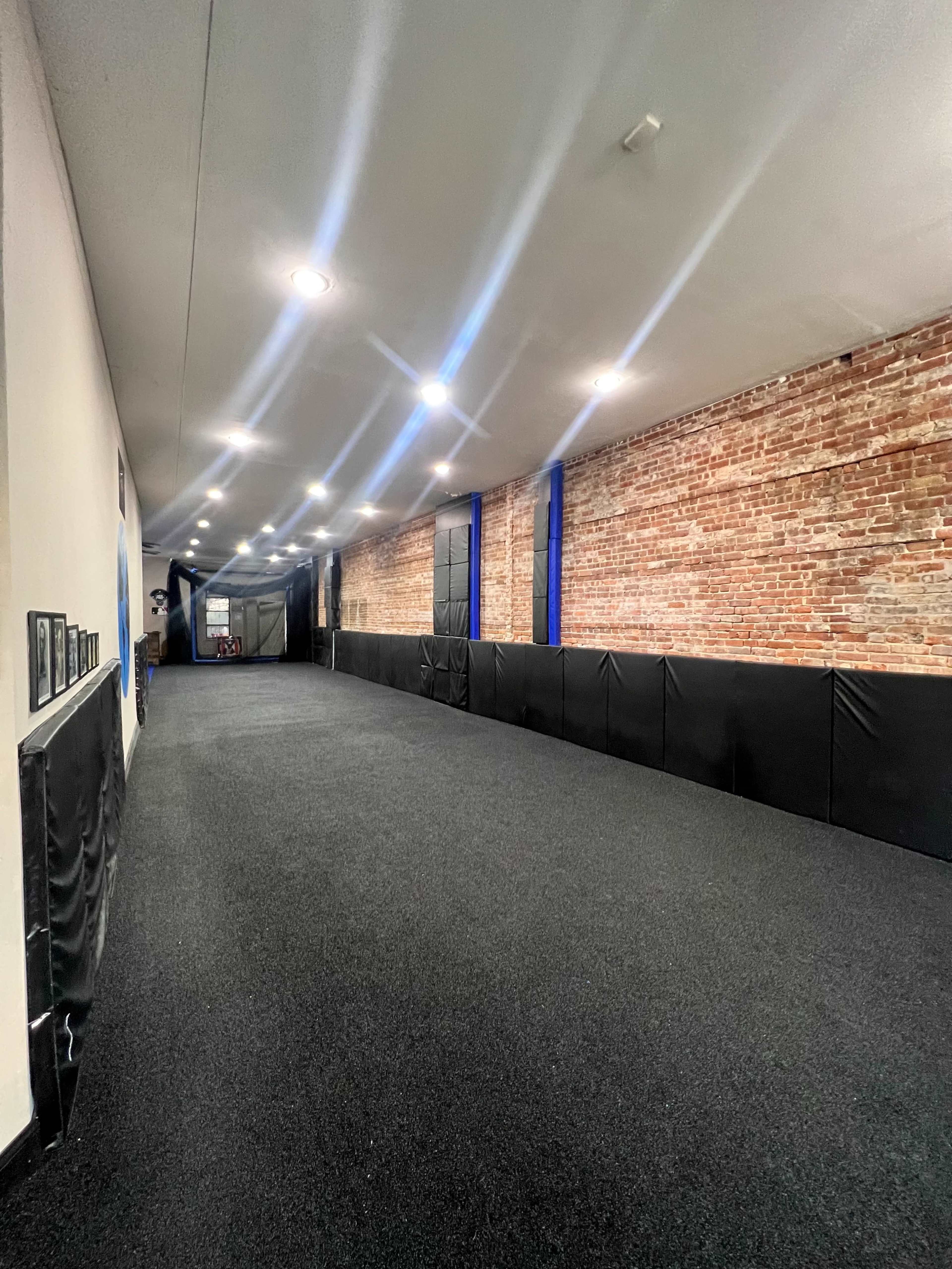 A long, narrow training space with a dark carpeted floor and exposed brick walls, illuminated by overhead lights.
