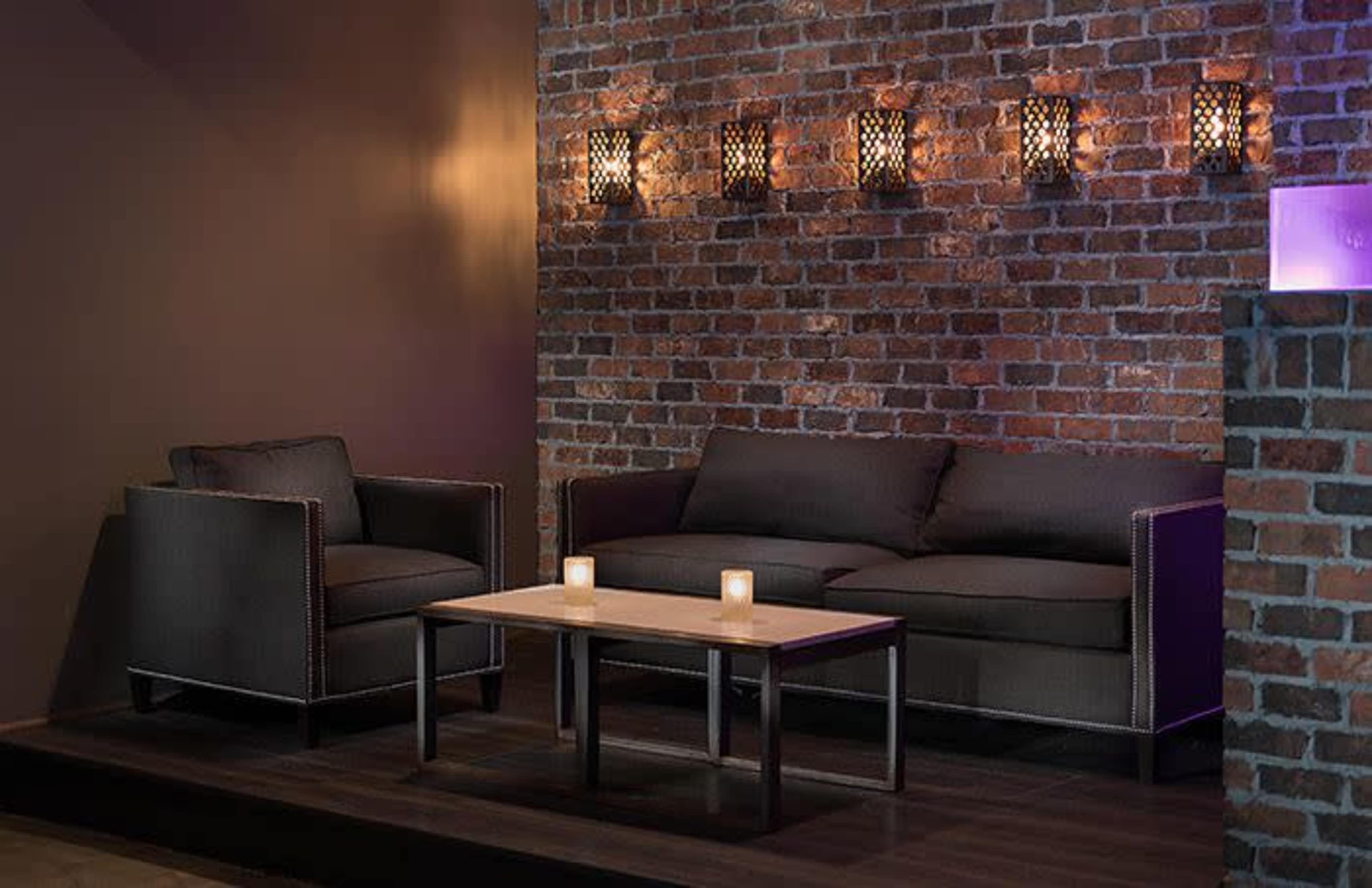 A modern seating area with a dark brick wall, a sofa, an armchair, a coffee table, and candles arranged on the table.