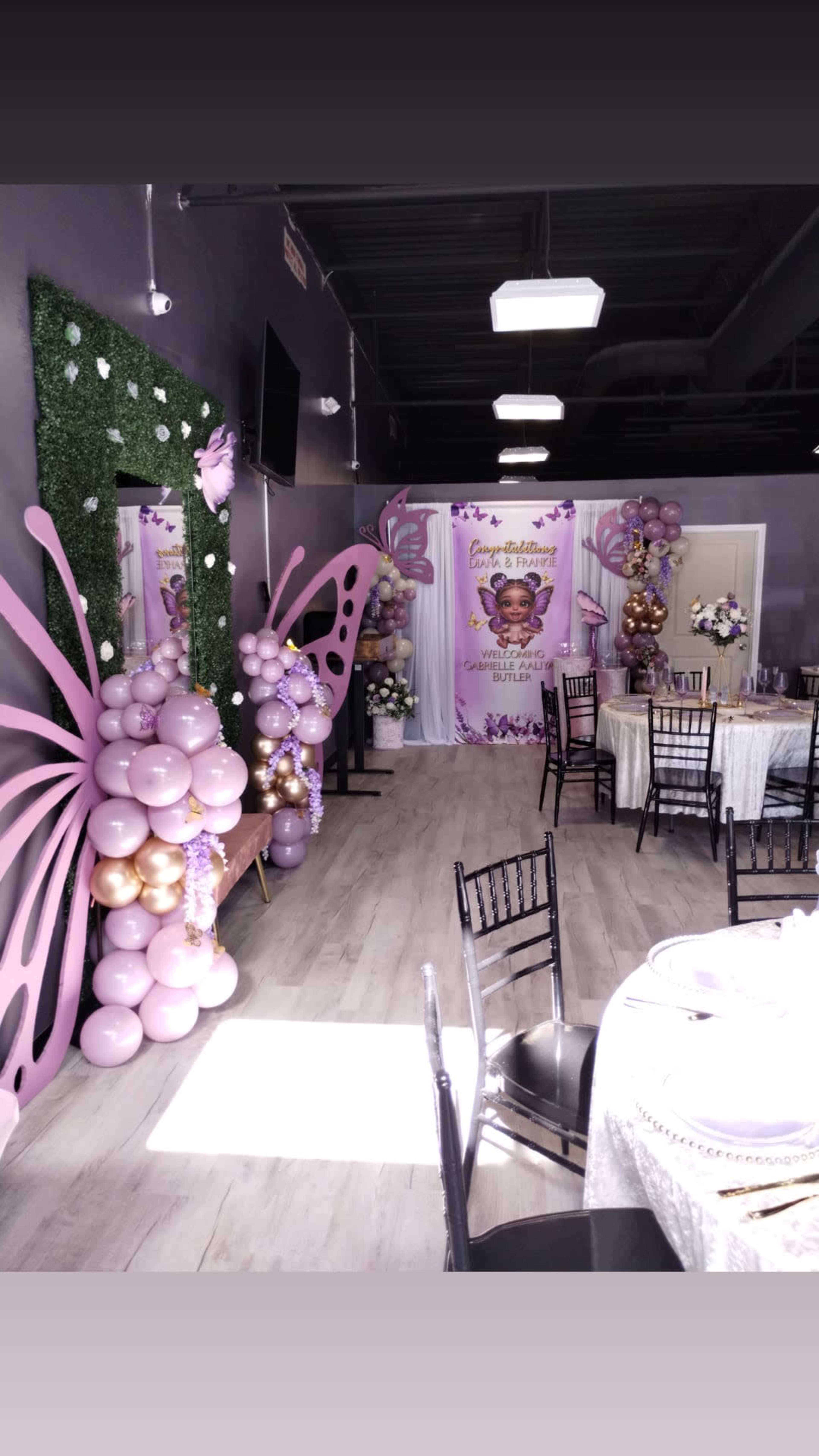 The image shows a decorated event space with a butterfly theme, featuring purple and gold balloons, floral arrangements, and tables set for a celebration.