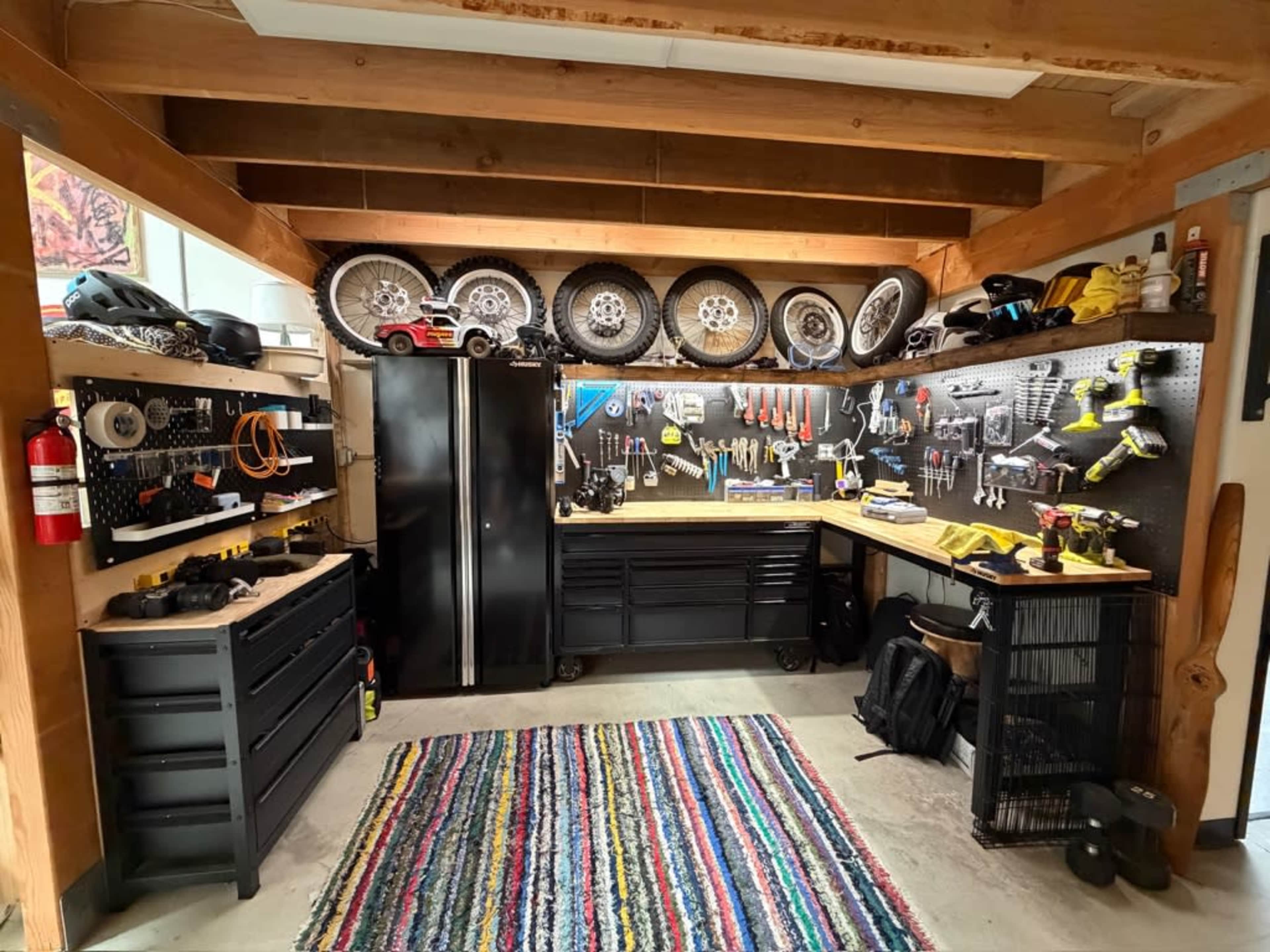 The image shows a neatly organized garage workshop featuring tools and equipment mounted on pegboards, two workbenches, and a colorful rug on the floor.