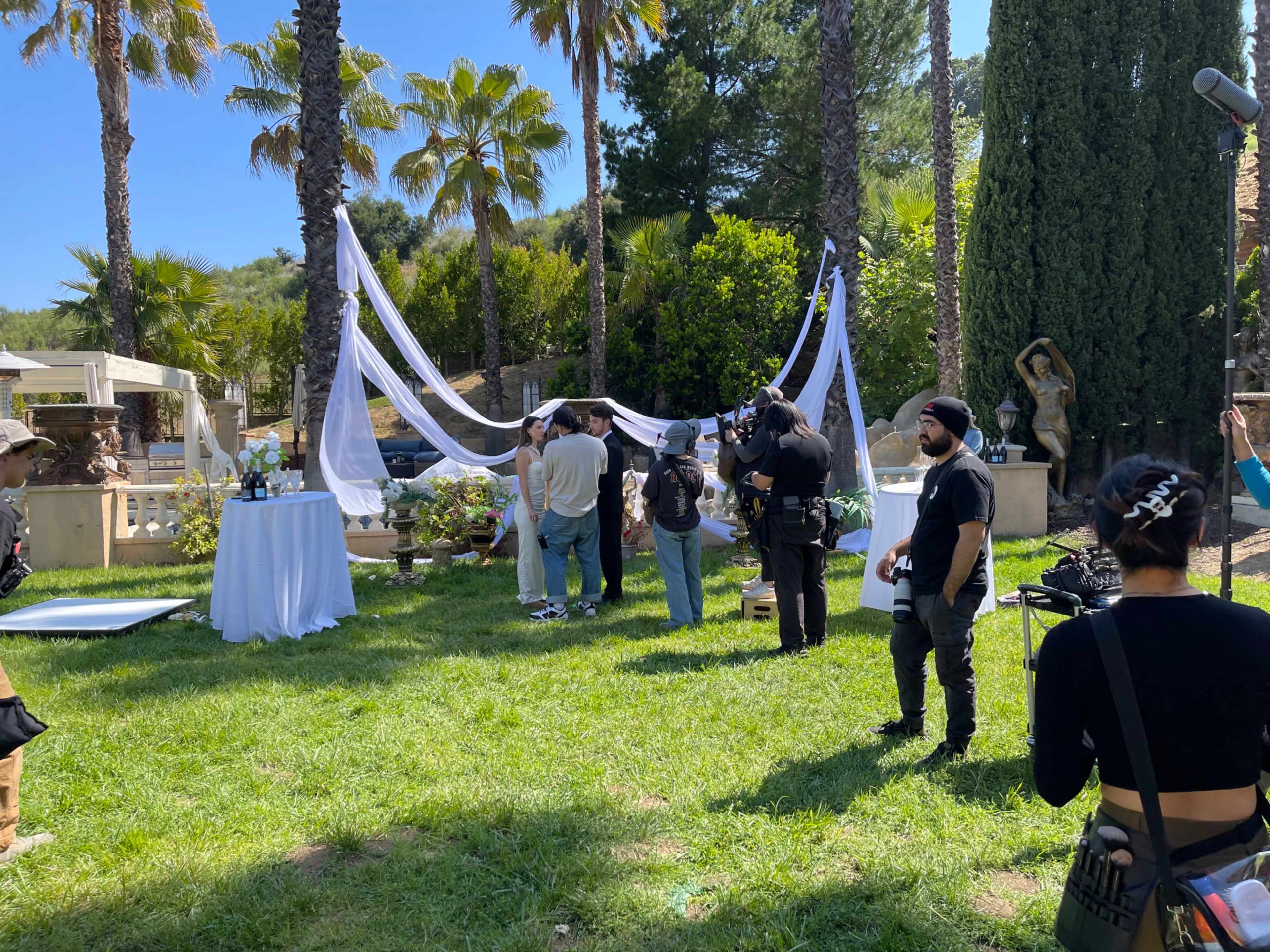 A group of people is gathered outdoors in a garden setting, with photographers capturing the scene among decorative drapery and tropical plants.