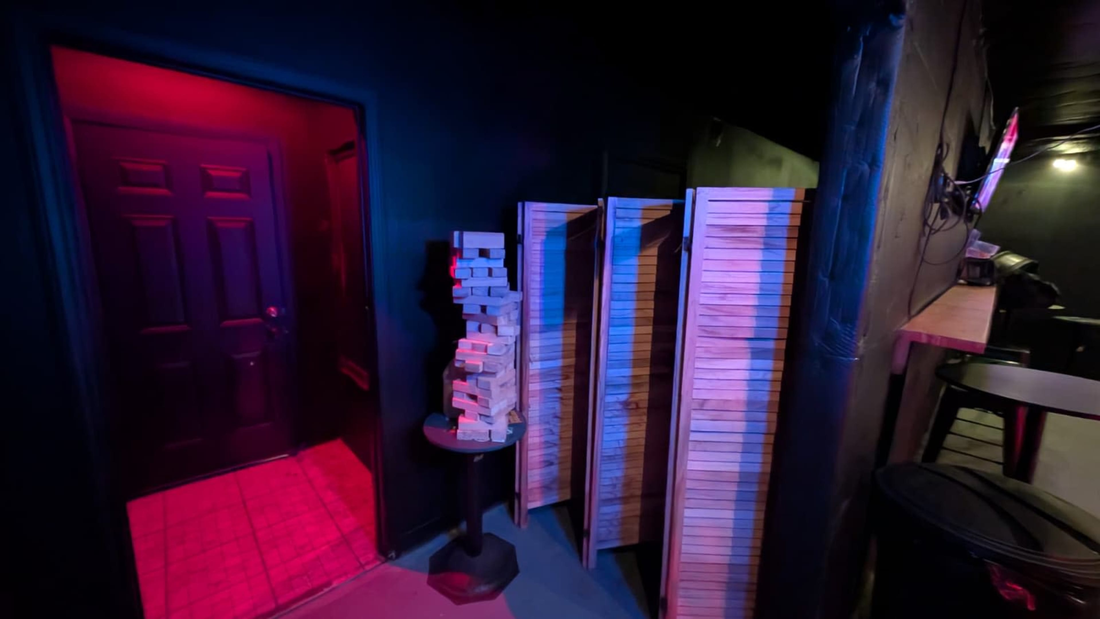 The image shows a dimly lit room with a dark wall, a door on the left emitting red light, and a stack of wooden blocks next to wooden shelves.