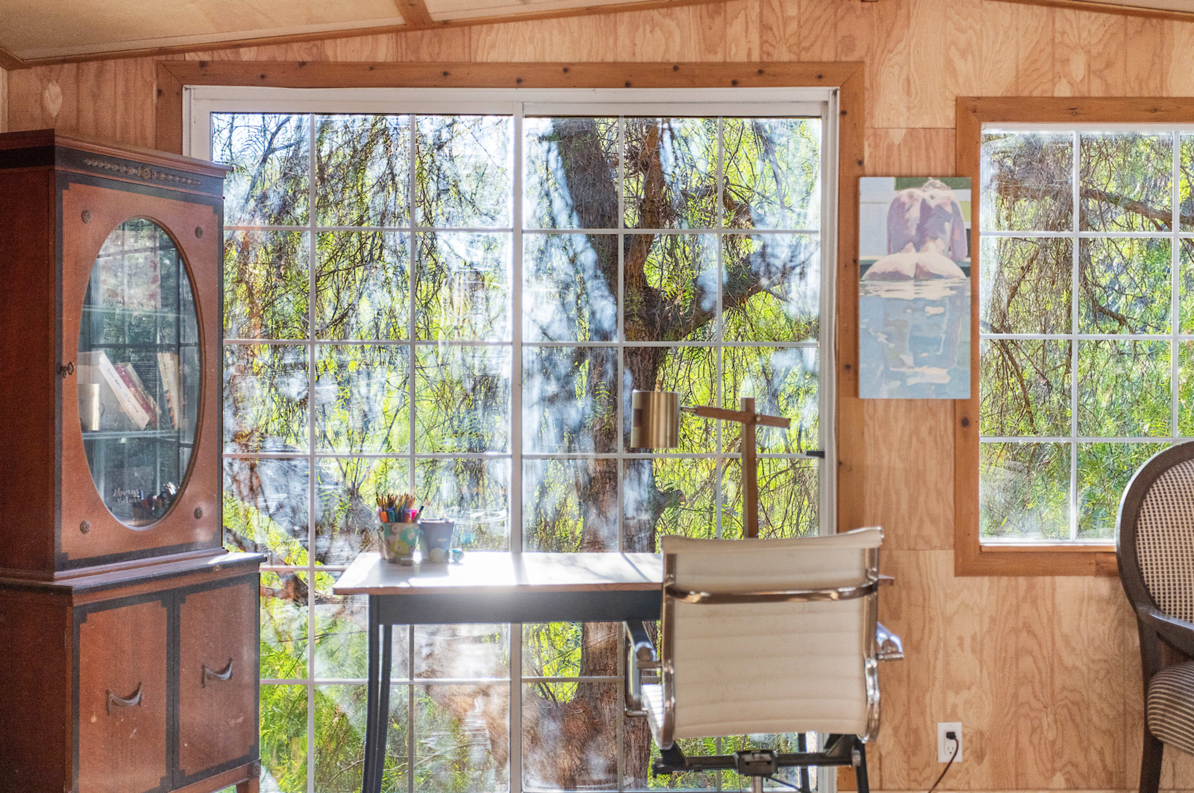 A cabin interior with a wooden wall, a desk and chair positioned beneath large windows that overlook greenery outside.