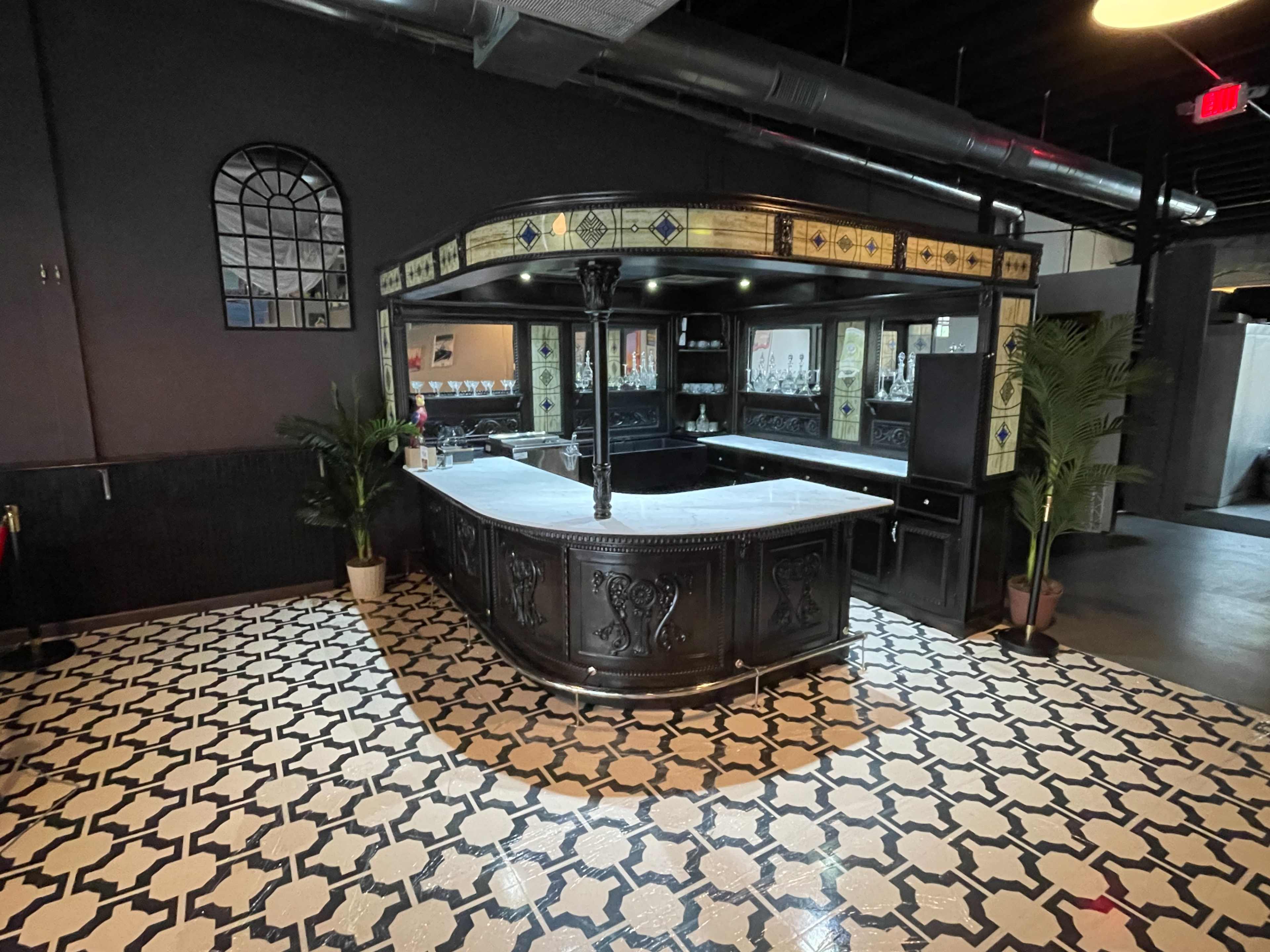 A black bar counter with a marble top, surrounded by patterned tile flooring and decorated with palm plants.
