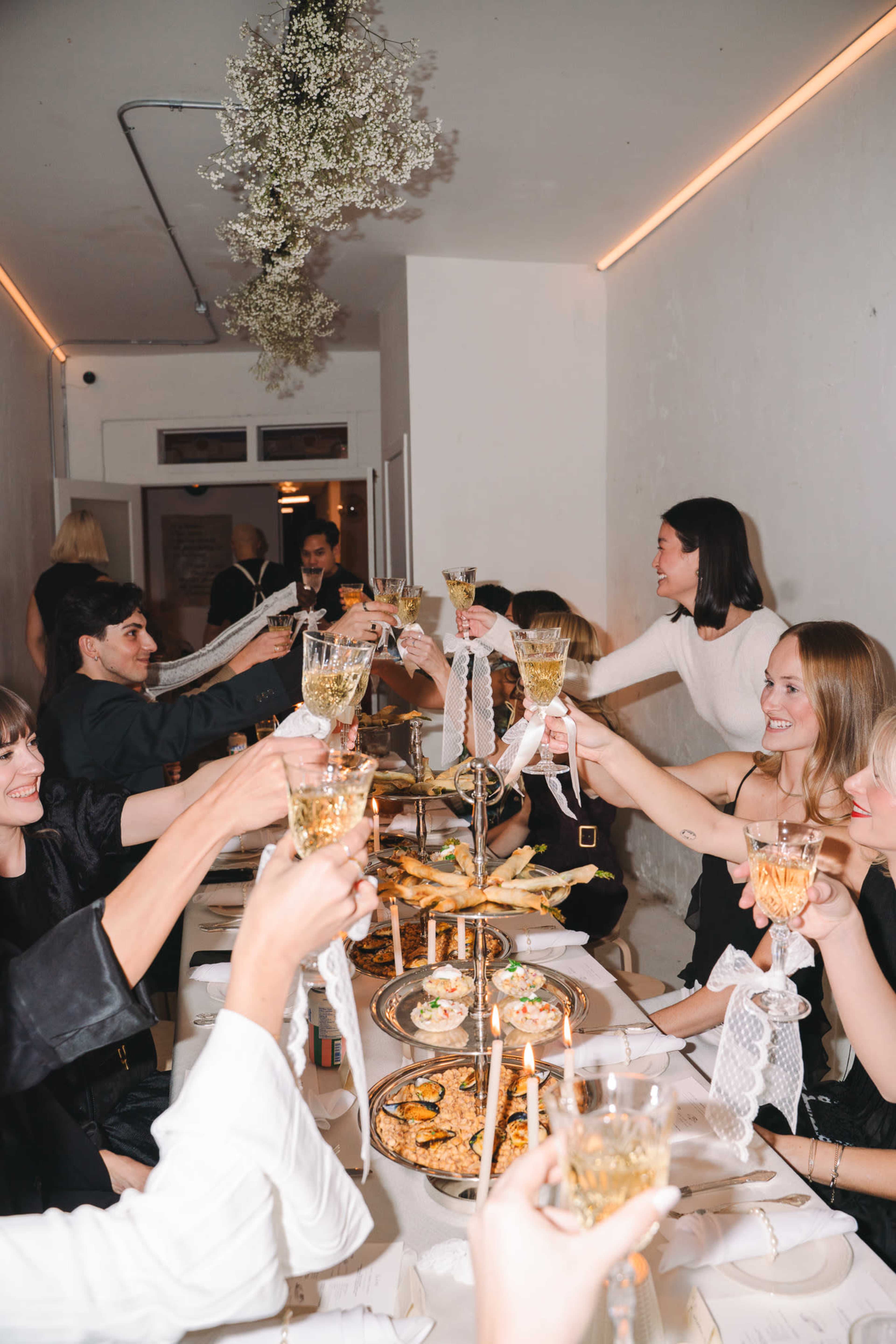 A group of people at a long table raise their glasses in a toast, surrounded by various dishes and decorations in a dimly lit setting.