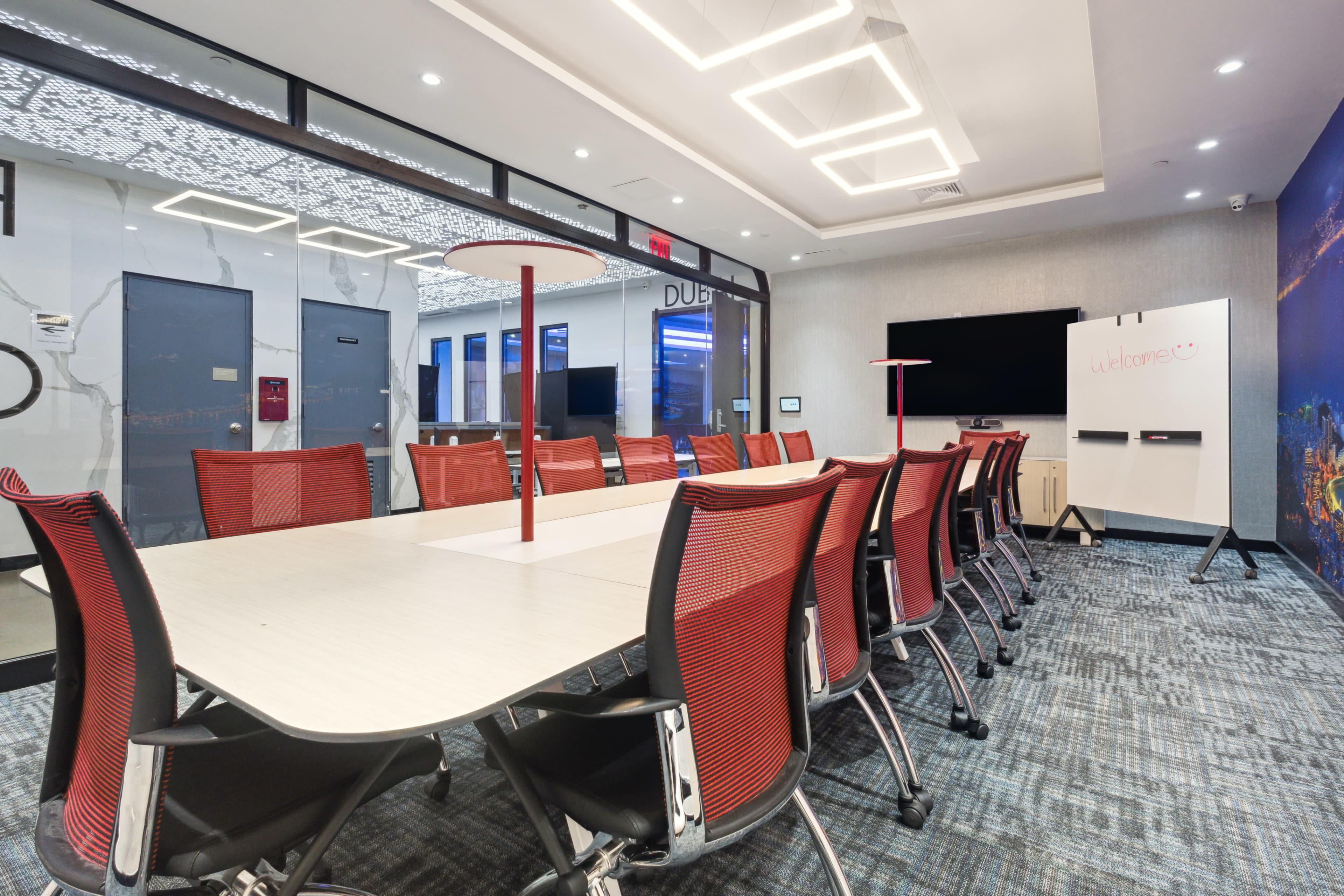 A modern conference room features a large table surrounded by red office chairs, with a whiteboard and a monitor on the wall.