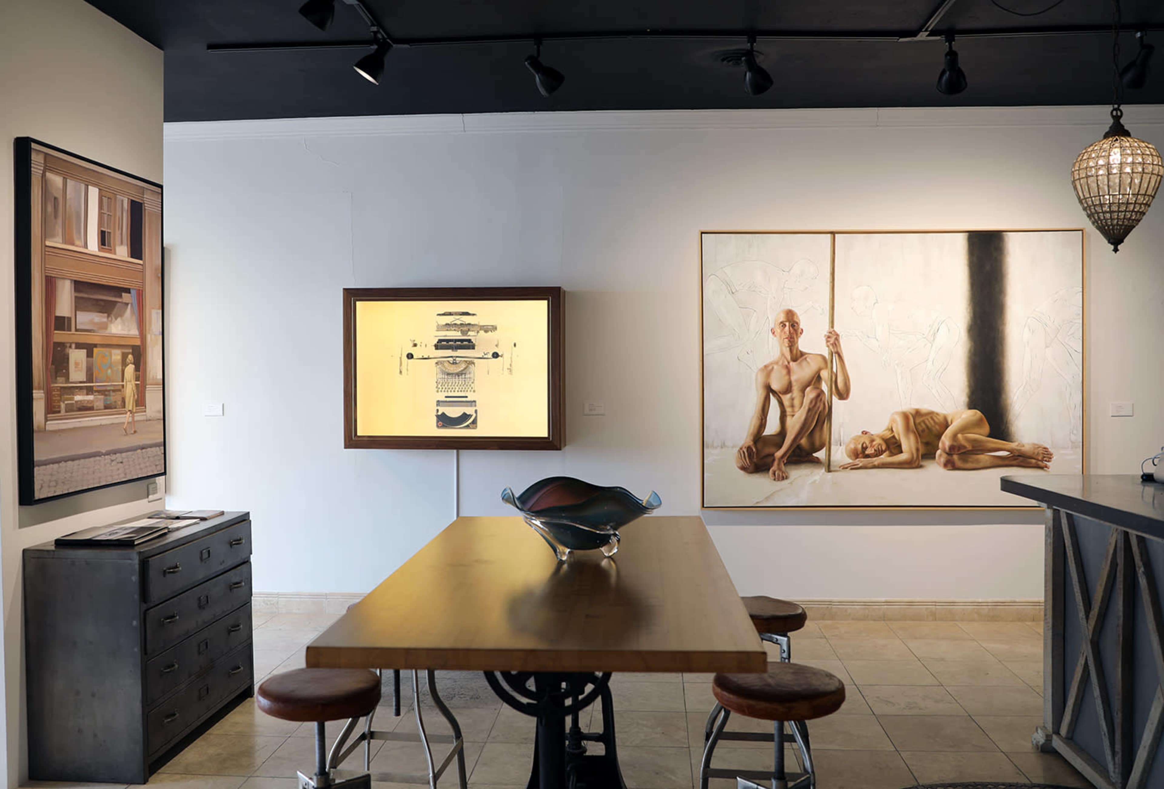 The image shows an art gallery with a wooden table in the center, surrounded by stools, and two artworks displayed on the walls.