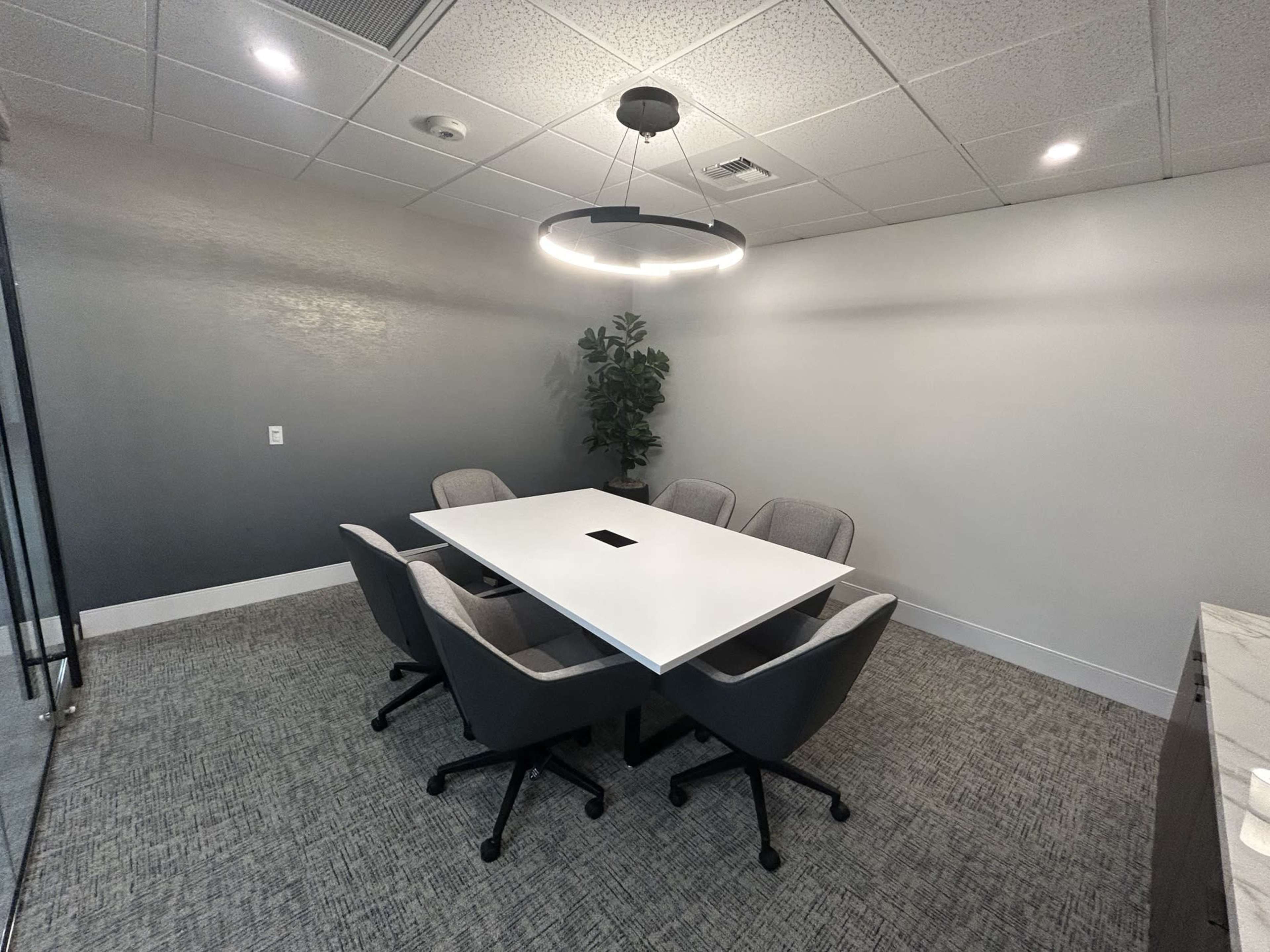 The image shows a modern conference room featuring a large white table surrounded by four gray chairs, with a plant and a circular light fixture overhead.