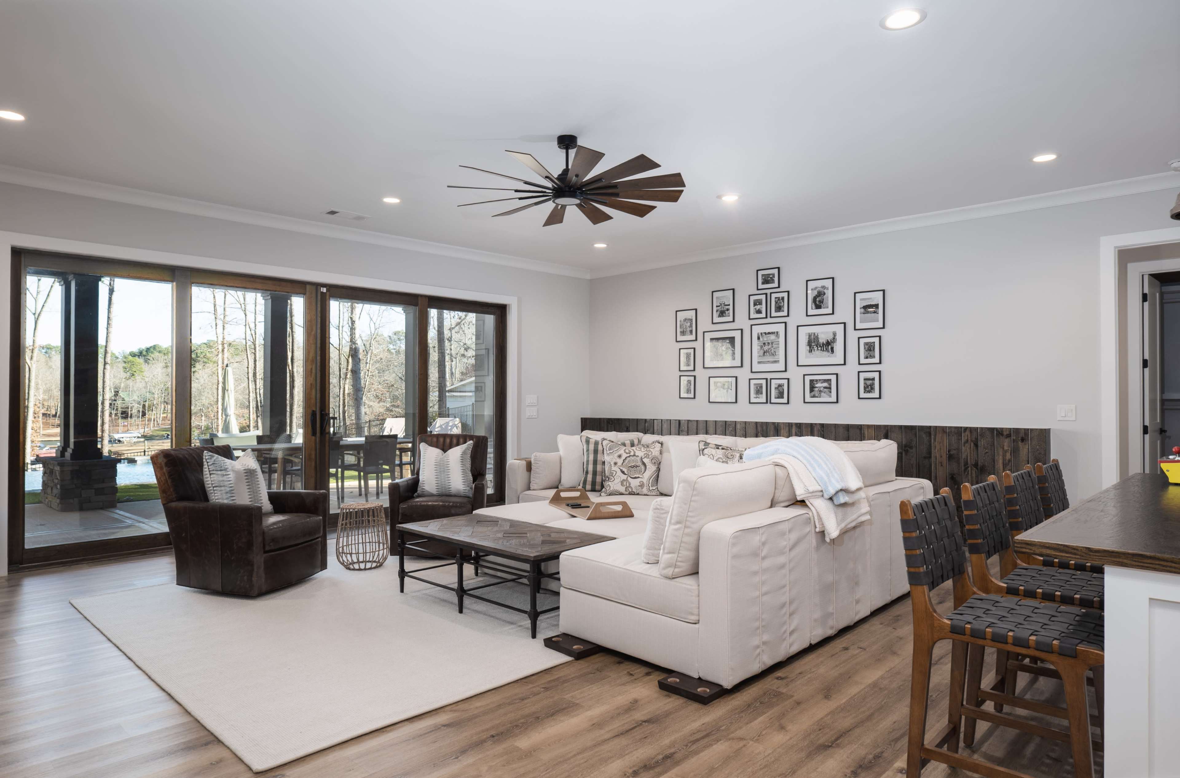A modern living room features a beige sectional sofa, dark wooden chairs, a large coffee table, and a gallery wall of framed photos, with large sliding glass doors providing a view of the outdoors.