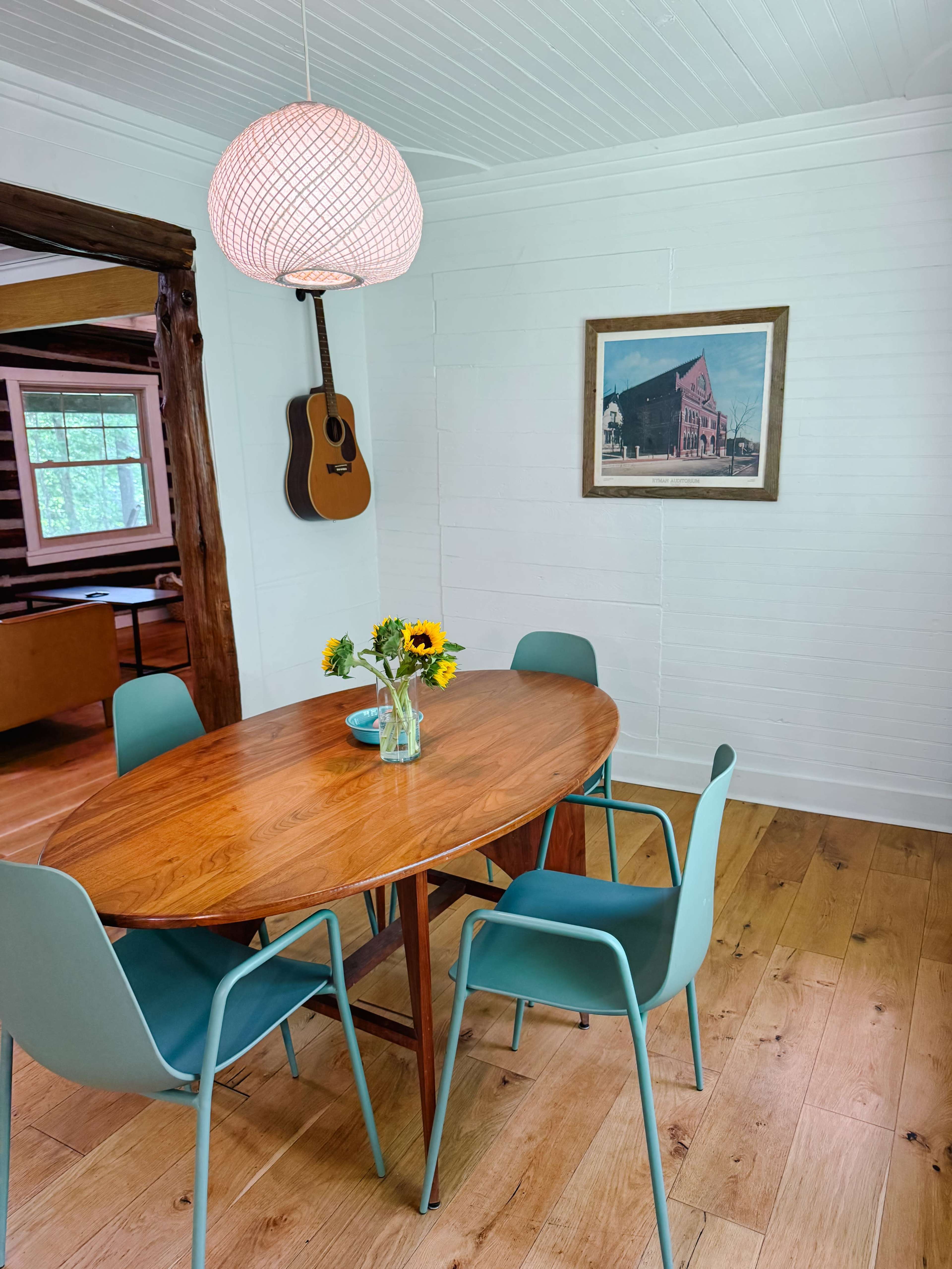 A wooden oval dining table with a vase of sunflowers is surrounded by four blue chairs under a hanging light fixture in a bright room with rustic decor.