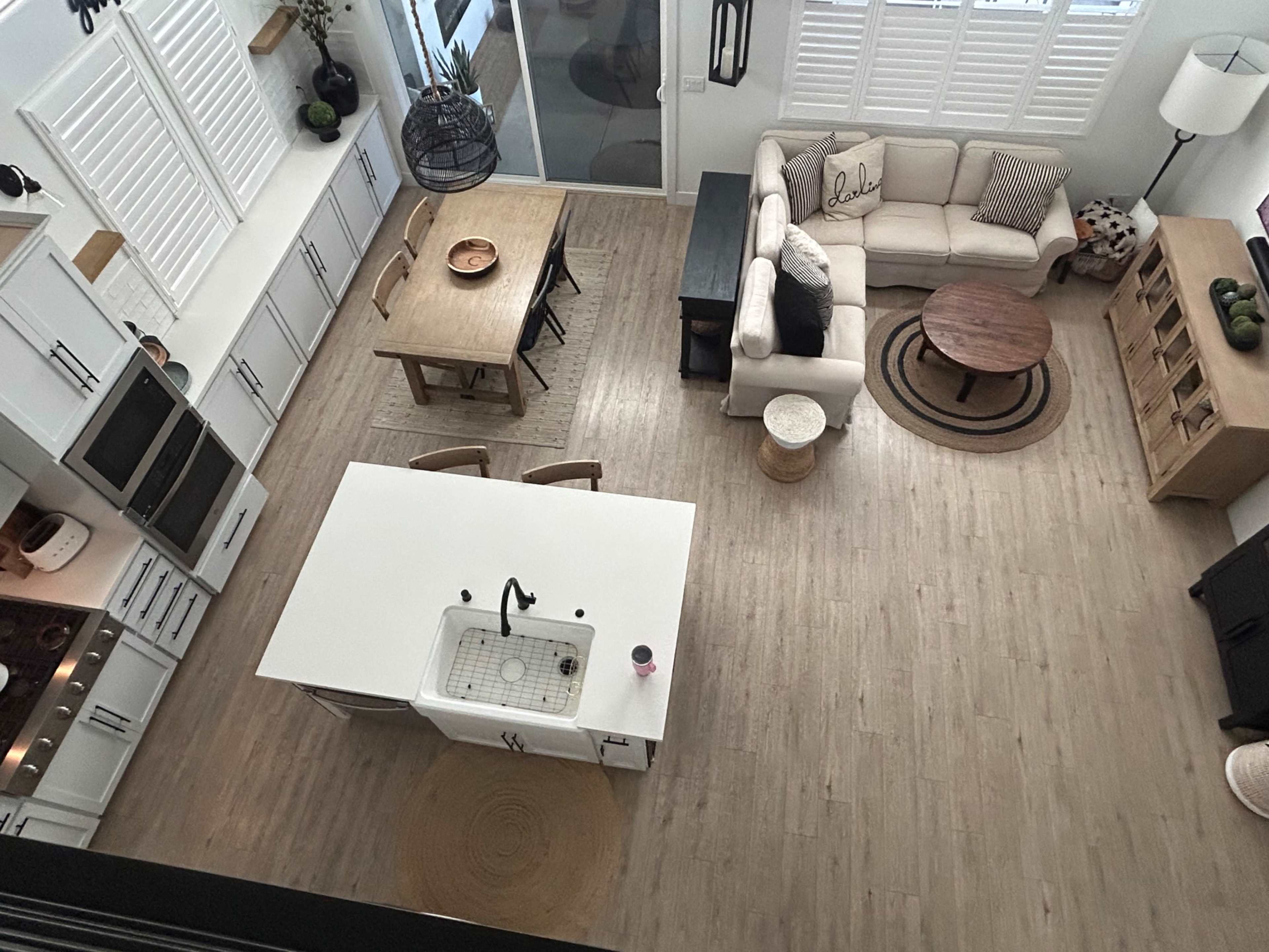 The image shows an overhead view of a modern kitchen and living area, featuring an island with a sink, a dining table, and a sectional sofa arranged around a coffee table.