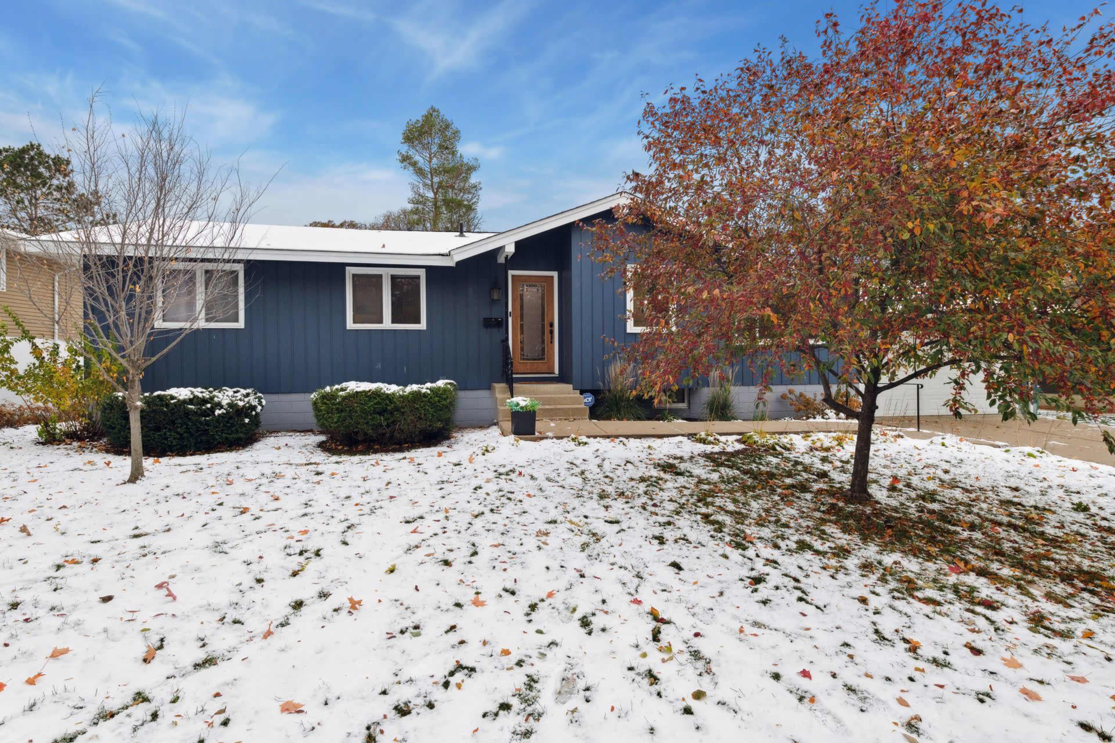 A blue house with a front door, surrounded by snow and a tree with red leaves.