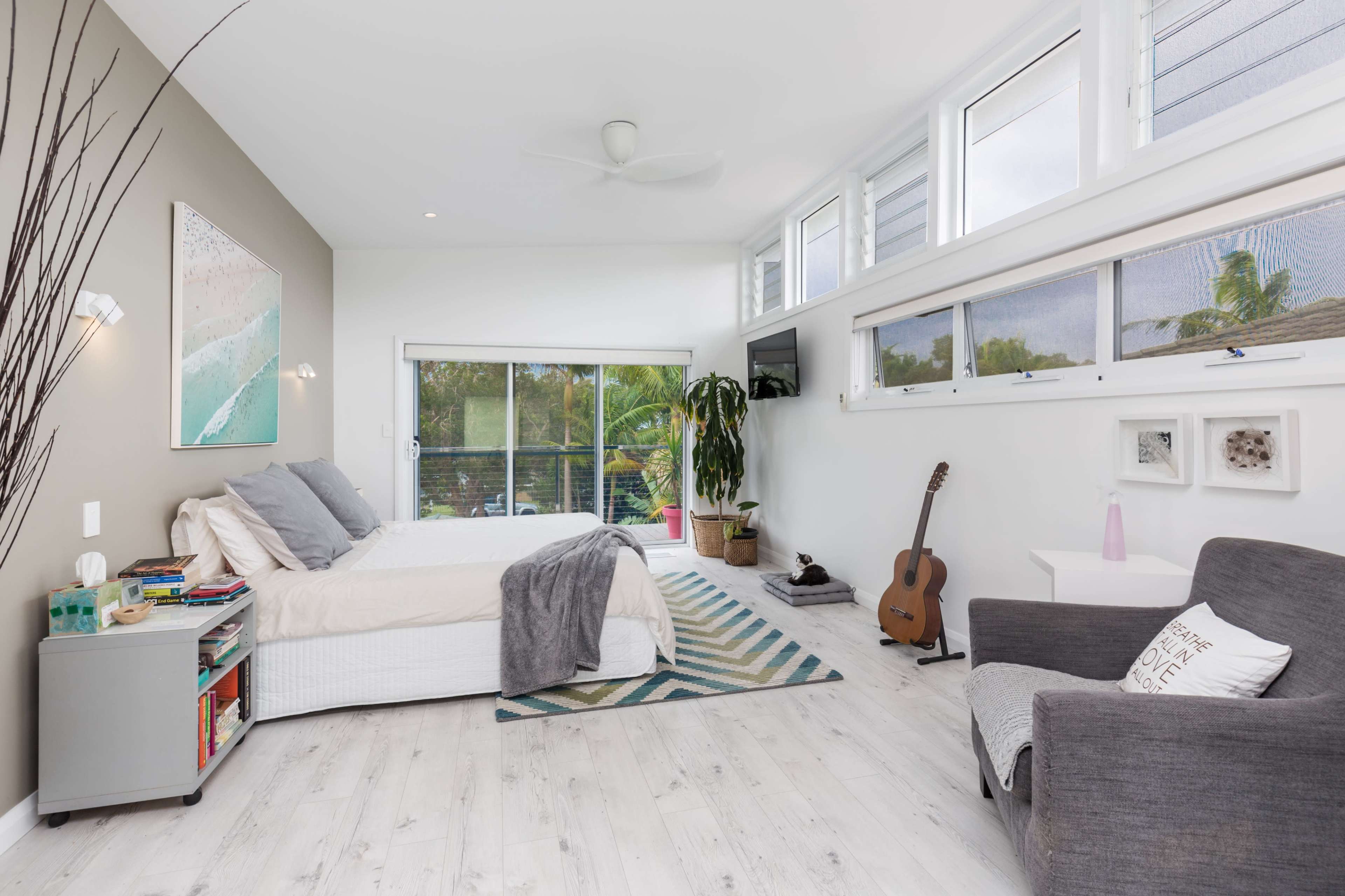 A modern bedroom features a bed with gray bedding, large windows, a potted plant, a guitar, and minimalist decor.