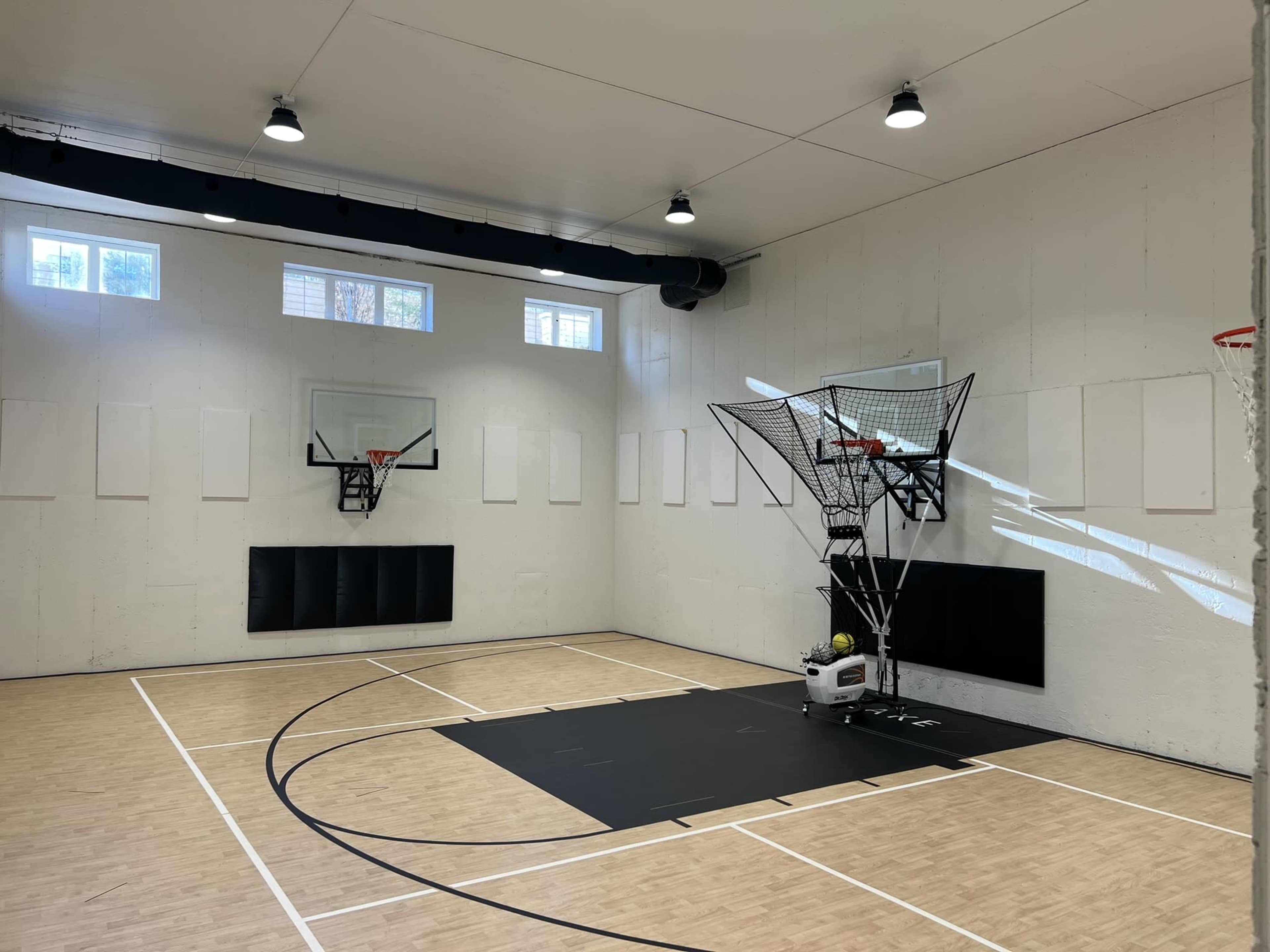 A modern indoor basketball gym featuring two hoops, a shooting machine, and a wooden floor.