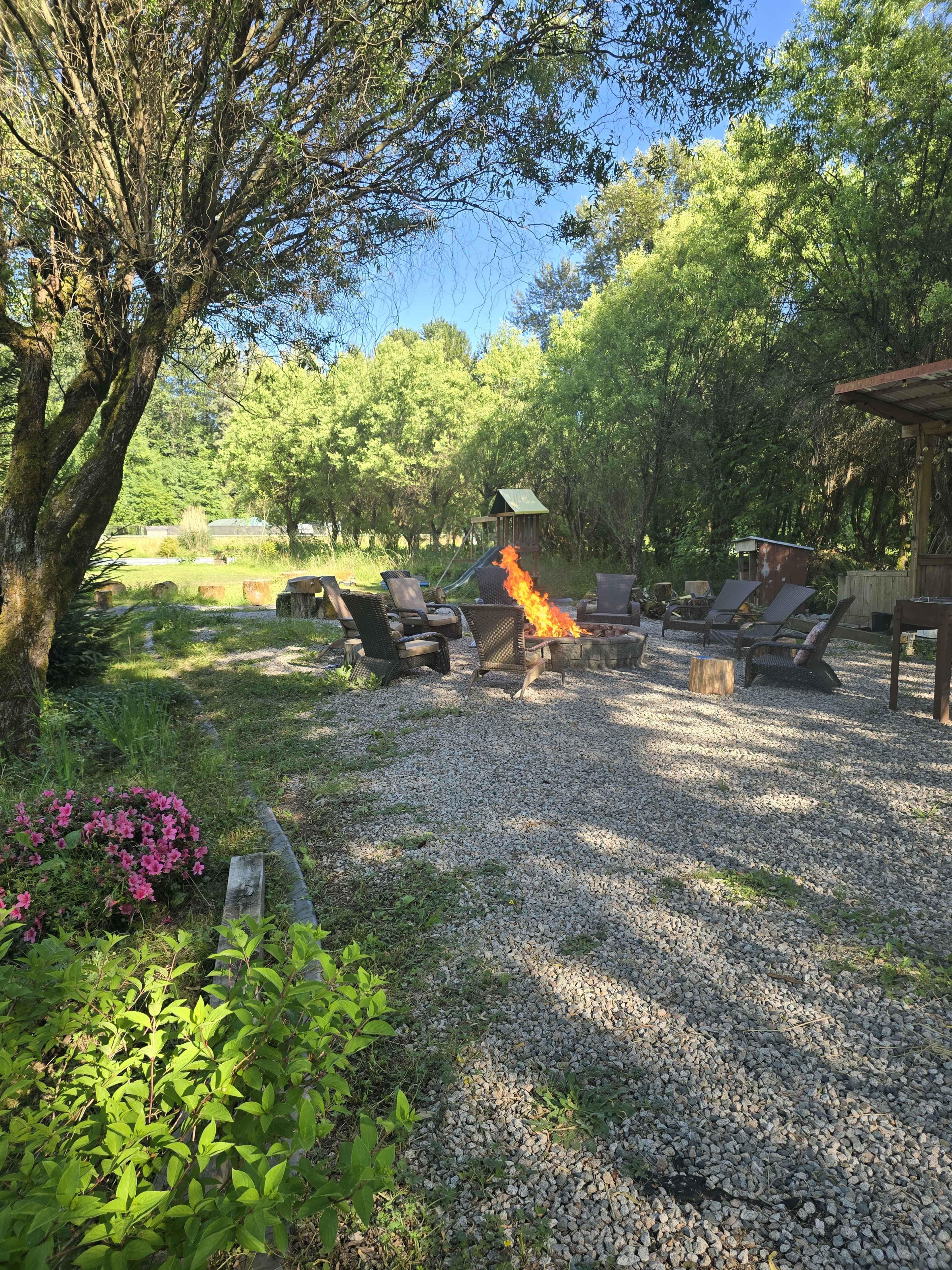 A fire burns in a metal fire pit surrounded by lounge chairs on a gravel patio, flanked by trees and colorful flowers.