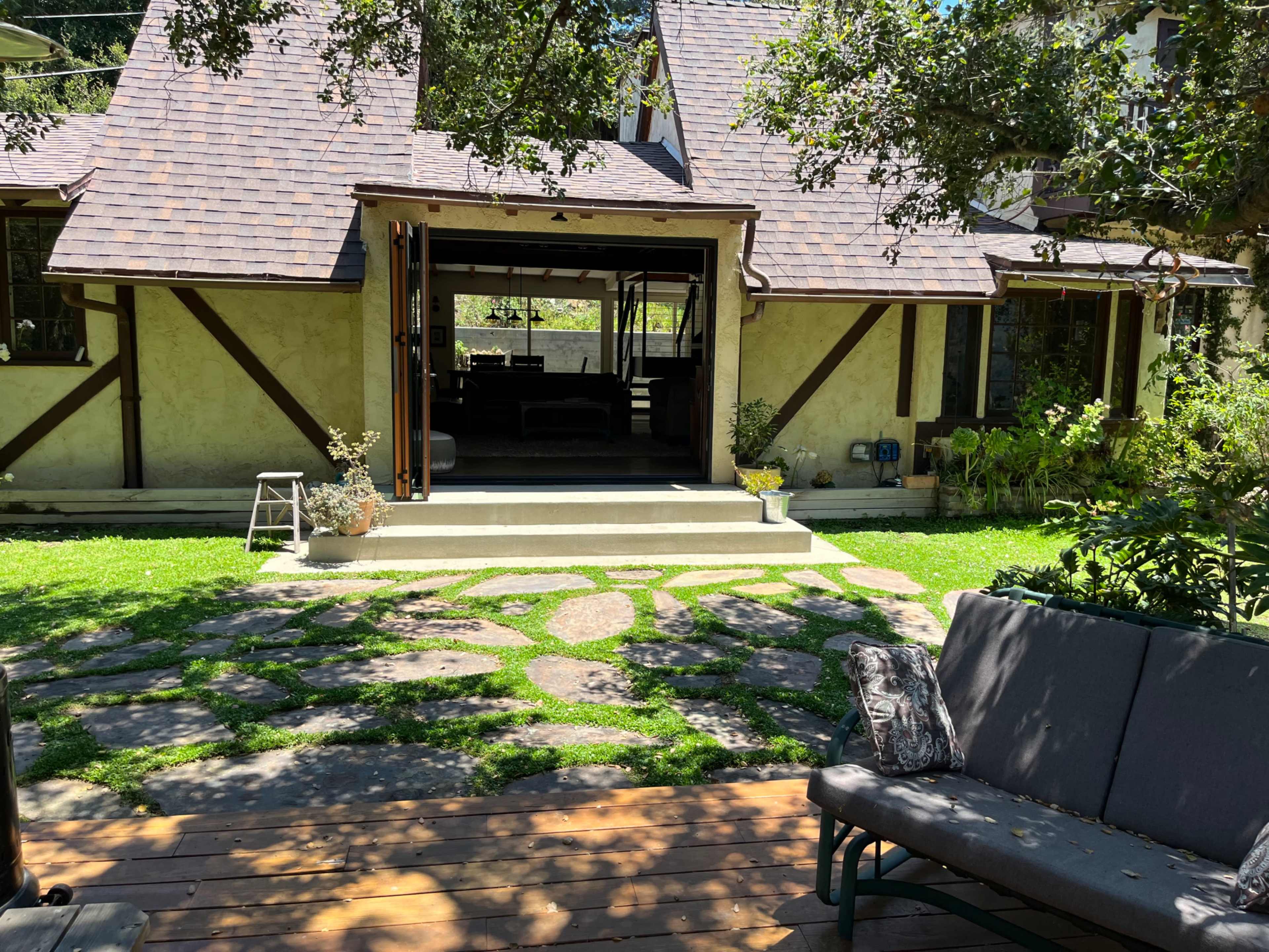 The image shows a backyard with a stone pathway leading to a house with a sloped roof and open doors, surrounded by greenery and outdoor seating.