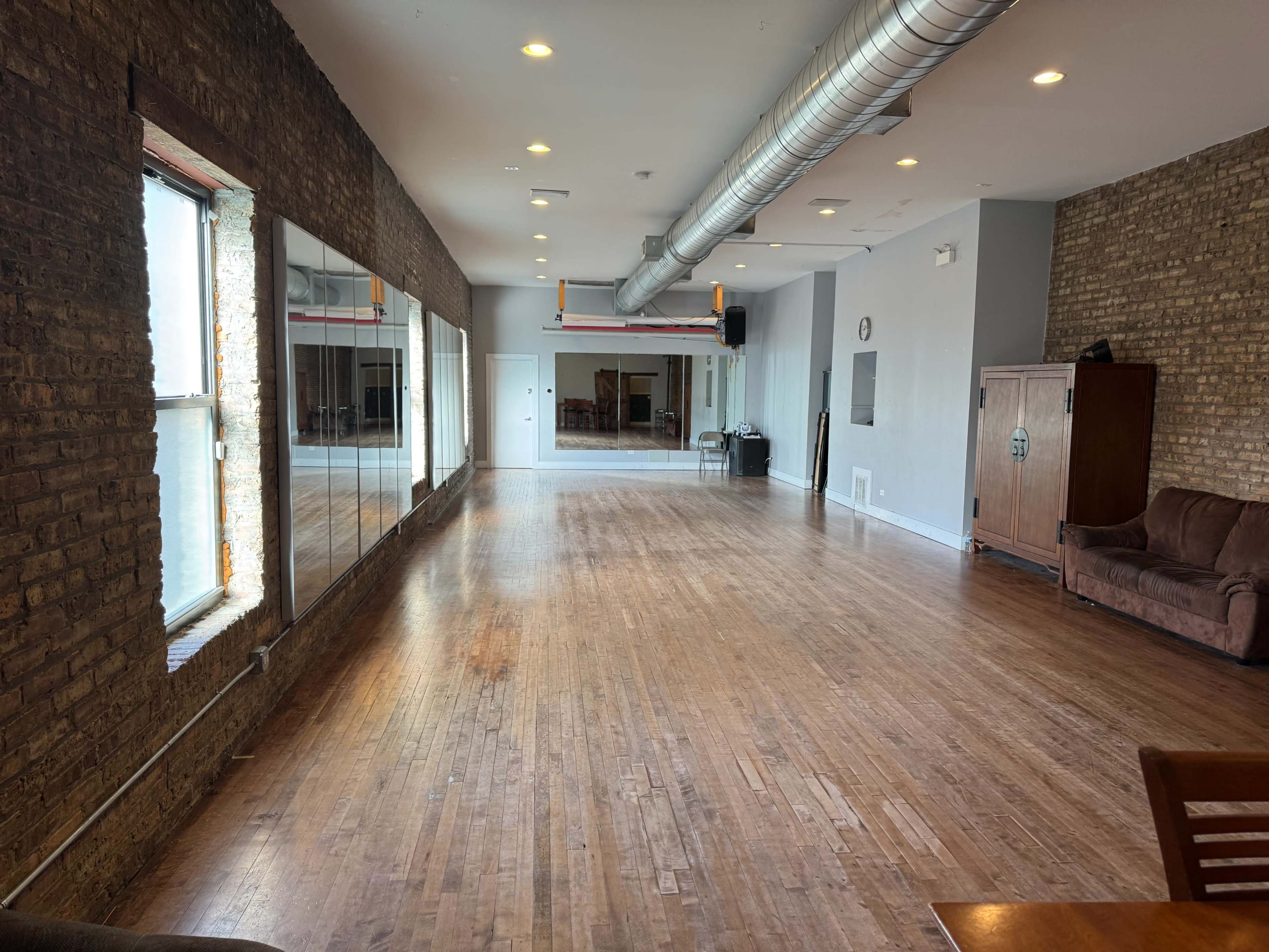 The image shows a spacious, empty room with wood flooring, large windows, and mirrored walls, accompanied by a few pieces of furniture along the sides.