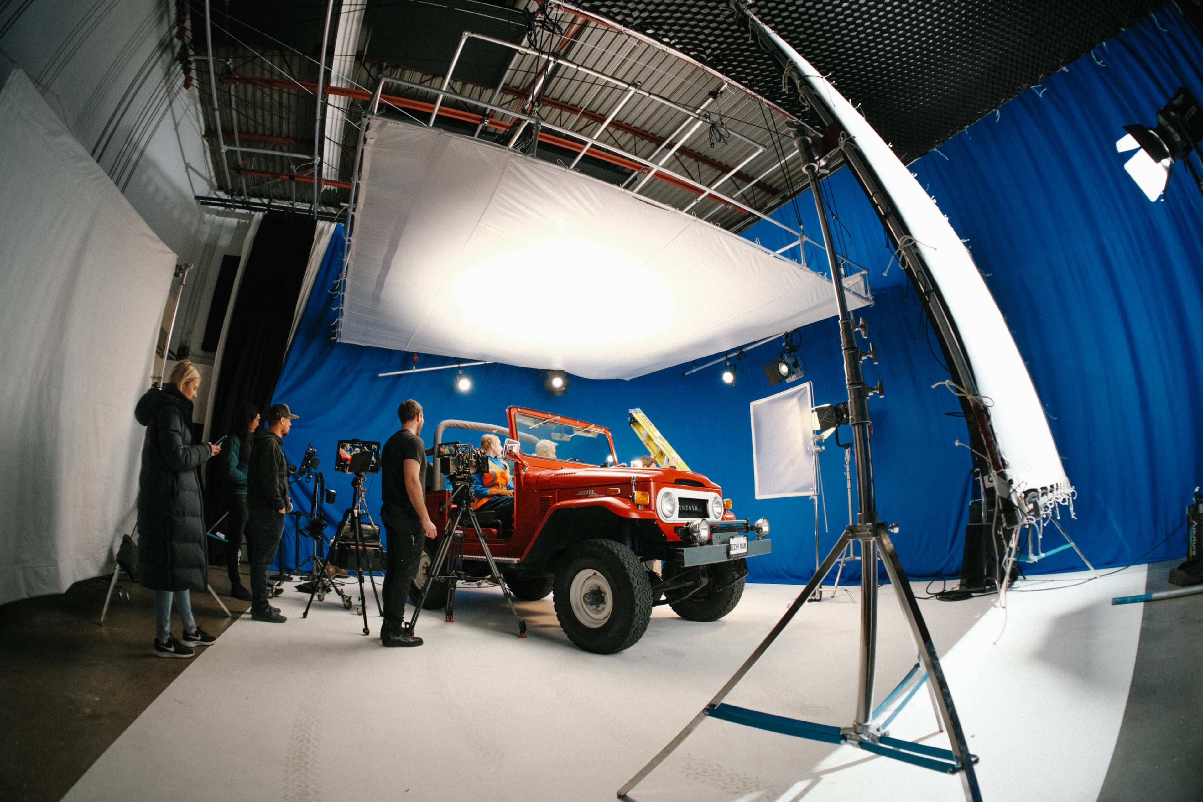 A vintage red jeep is being photographed in a studio setup with blue backdrops and professional lighting equipment.