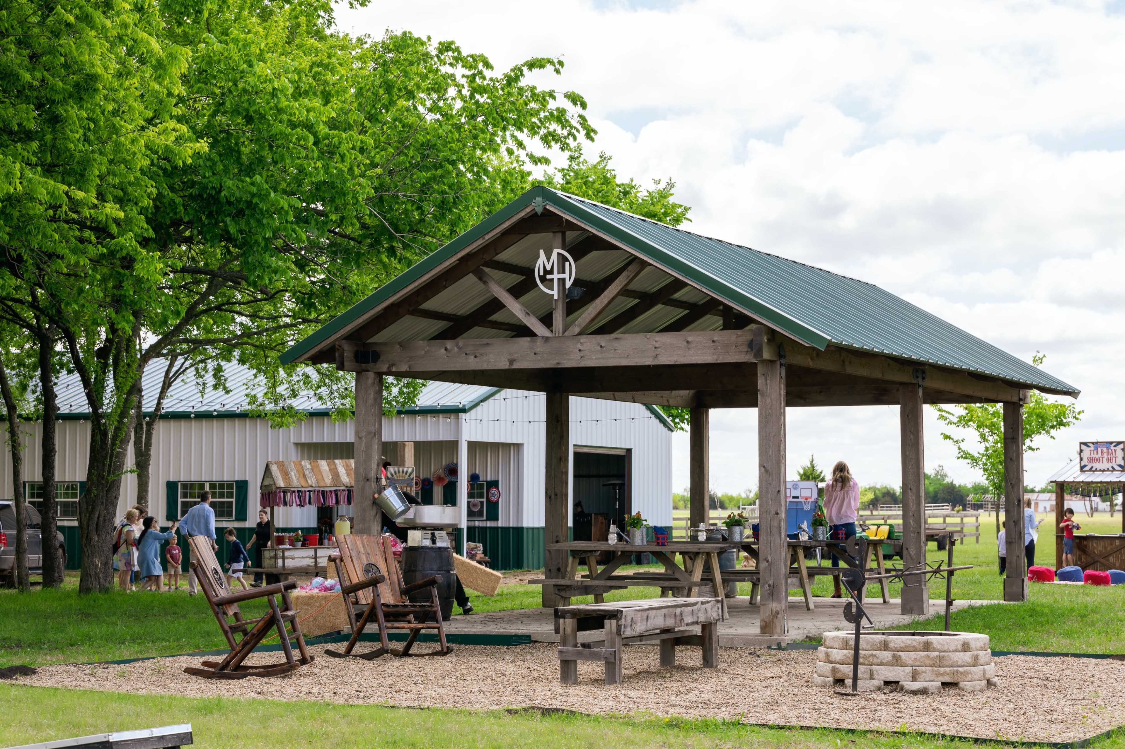 A shaded outdoor pavilion with wooden picnic tables and rocking chairs is set near a barn and grassy area.