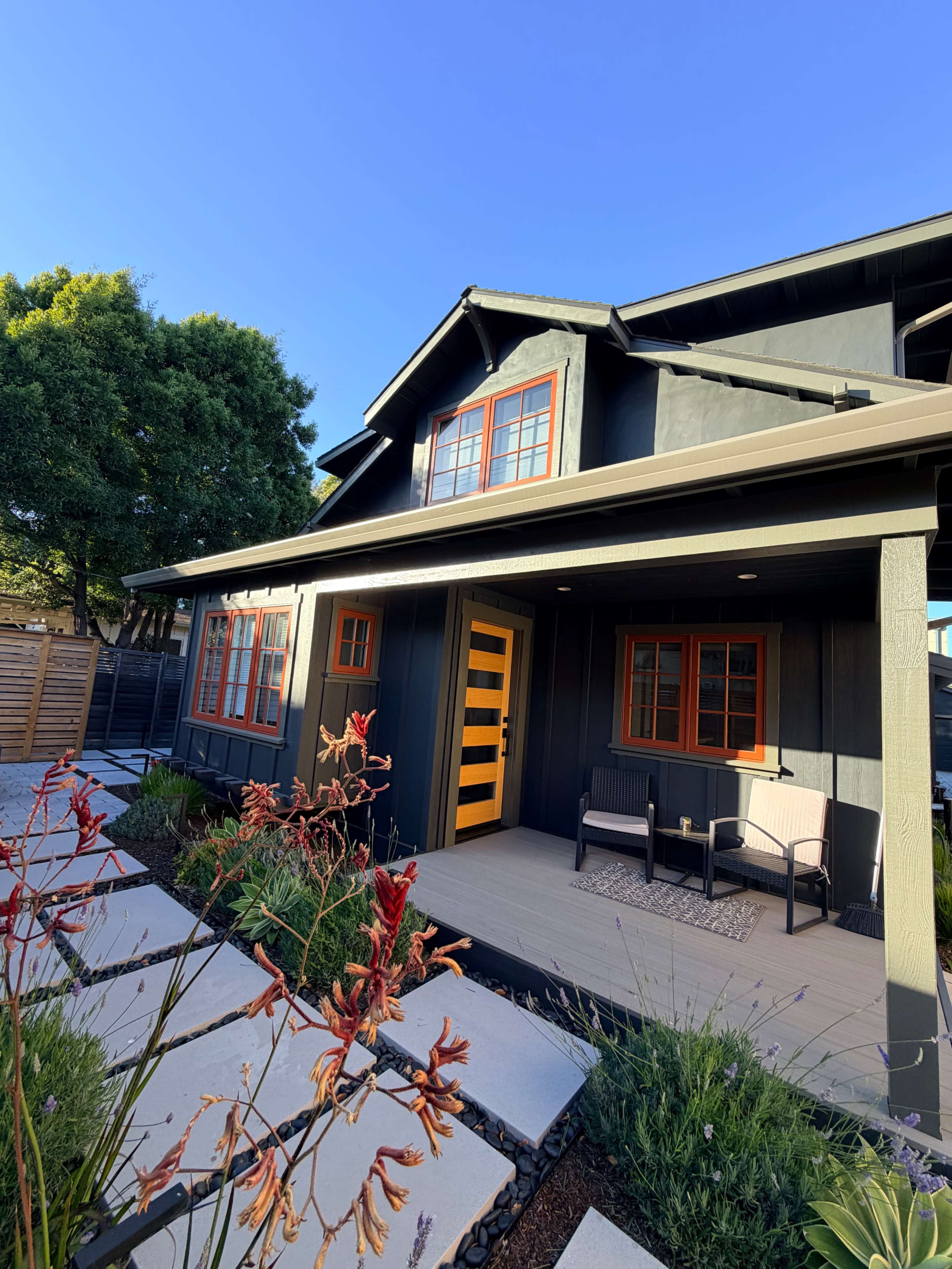 The image shows a modern black house with a striking yellow door, surrounded by landscaped plants and a small seating area on the porch.