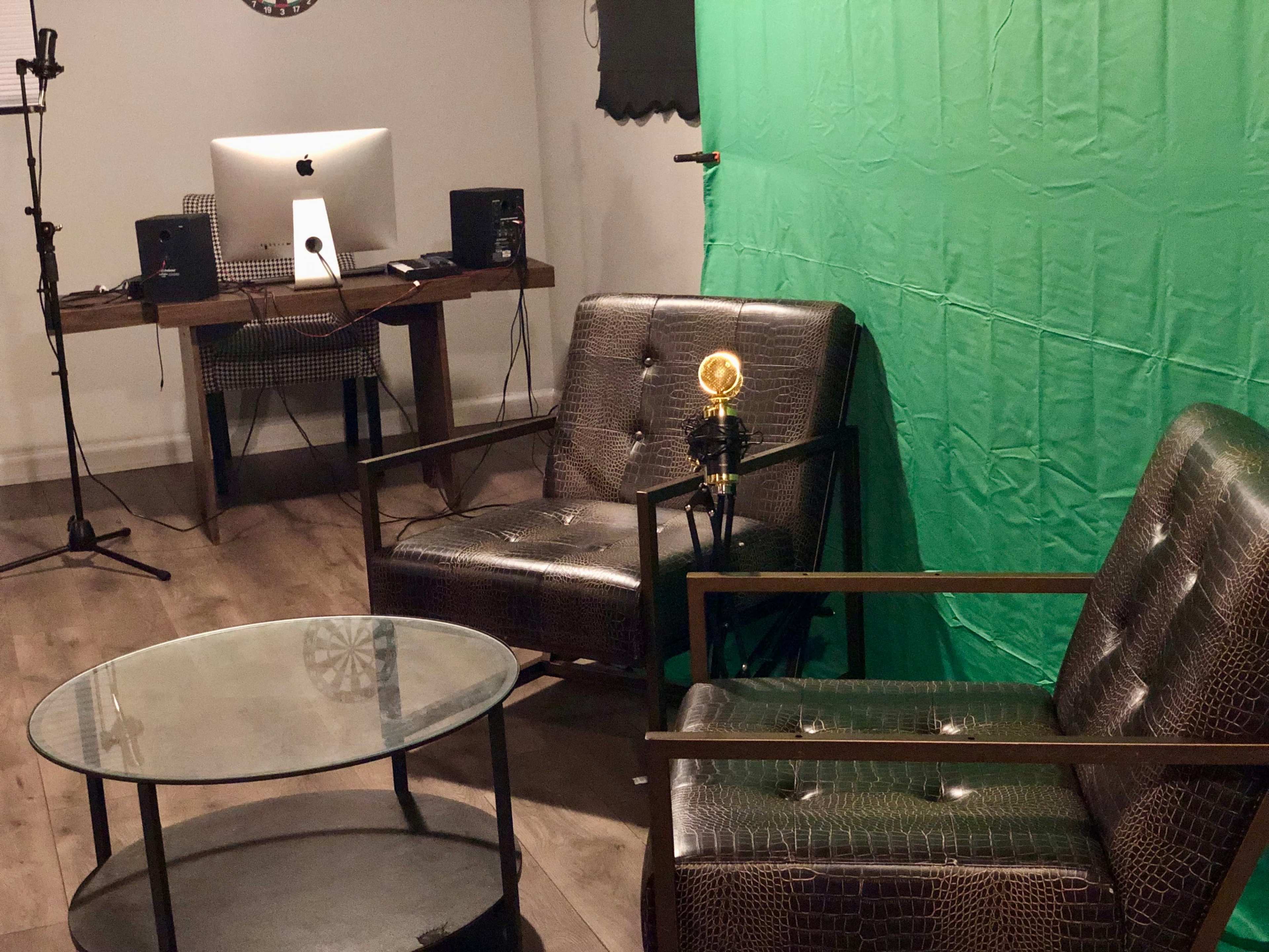 A room with a green screen backdrop, two patterned chairs, a glass coffee table, and a computer desk with an iMac setup.