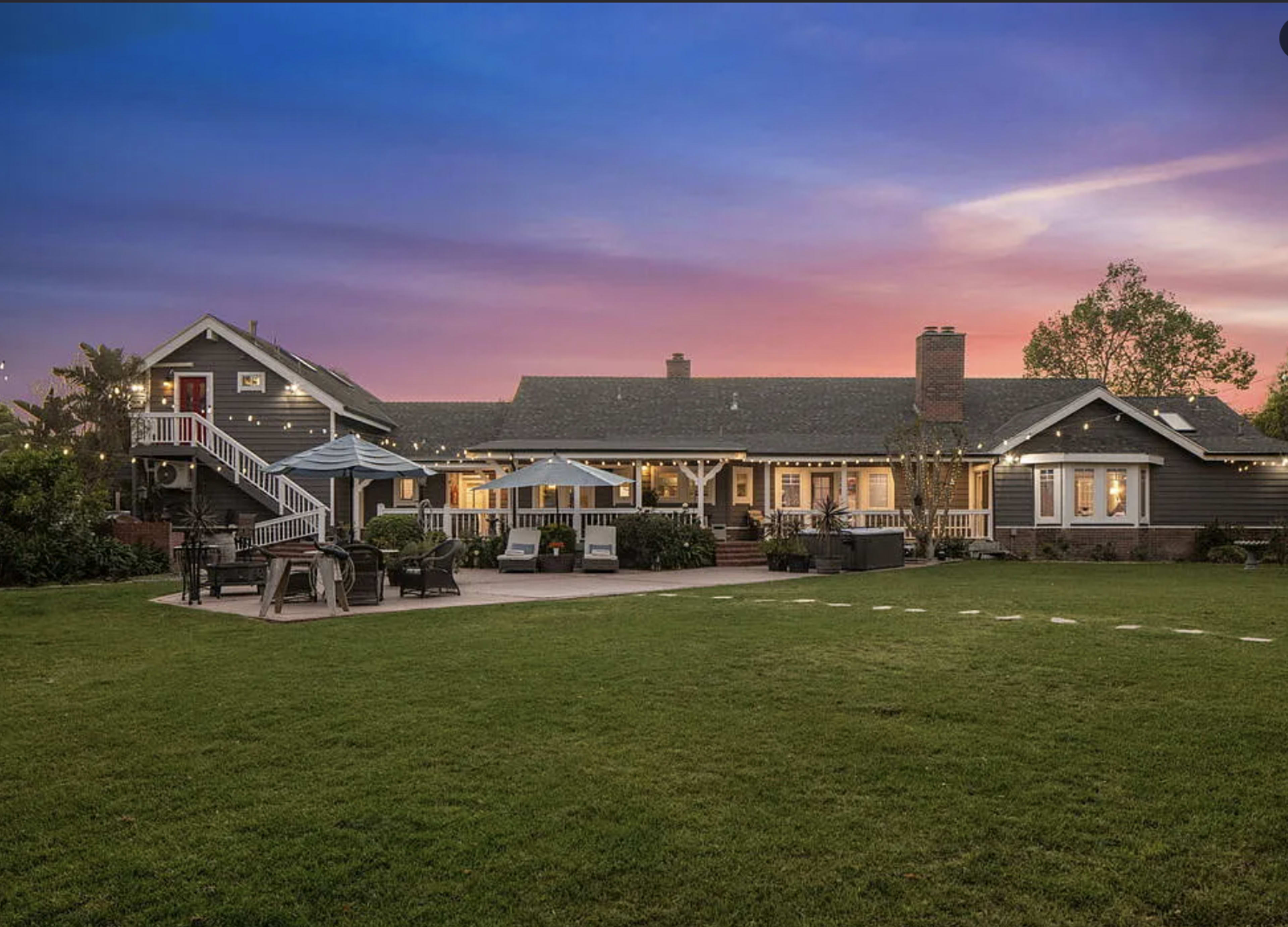 A two-story house with a large backyard and patio area is set against a colorful sunset sky.