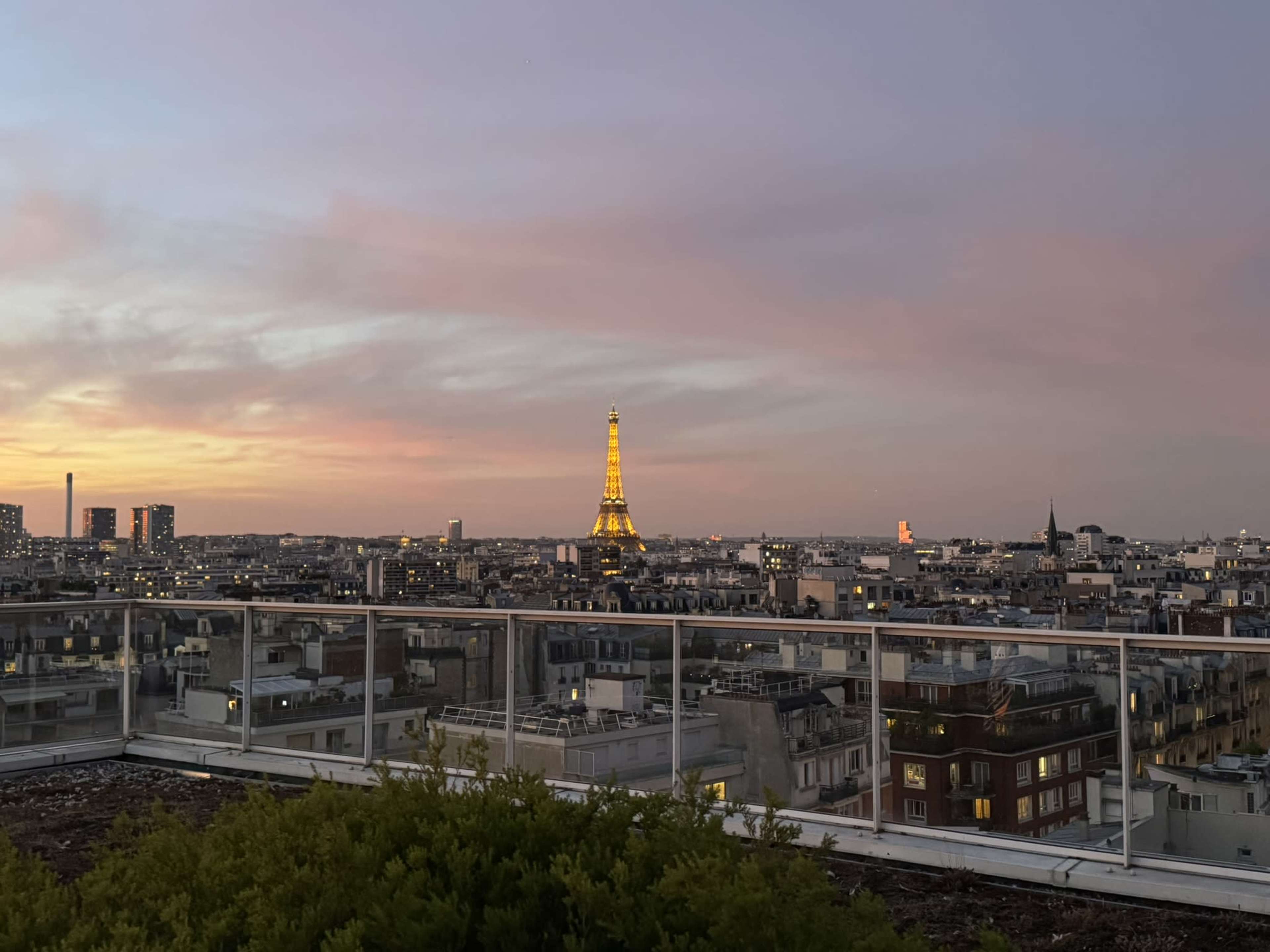 The image shows a panoramic view of Paris at sunset, highlighting the illuminated Eiffel Tower among the city's rooftops.