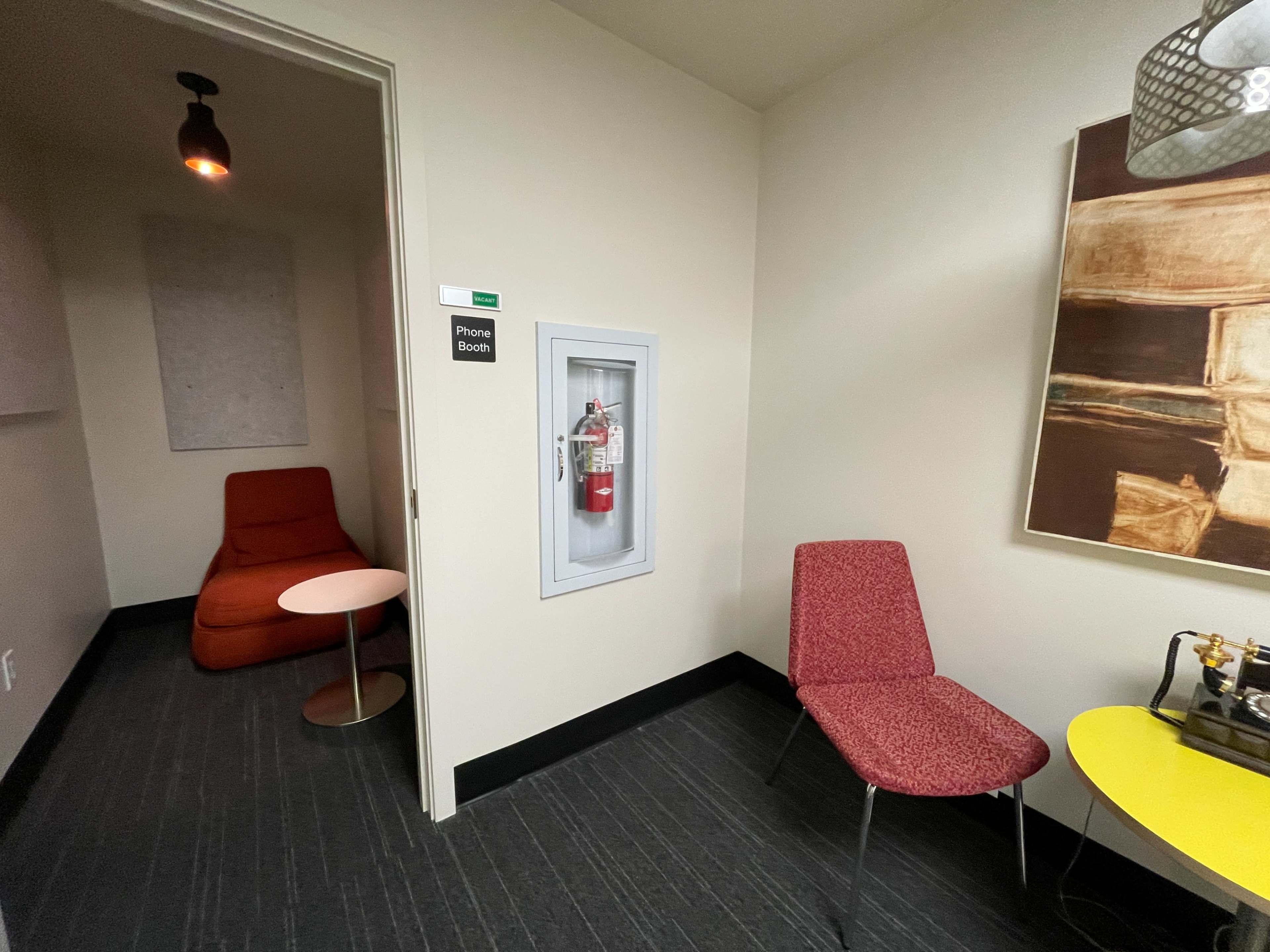 The image shows a small phone booth area featuring a red chair, a round table, a fire extinguisher on the wall, and a colorful yellow table beside a red chair.