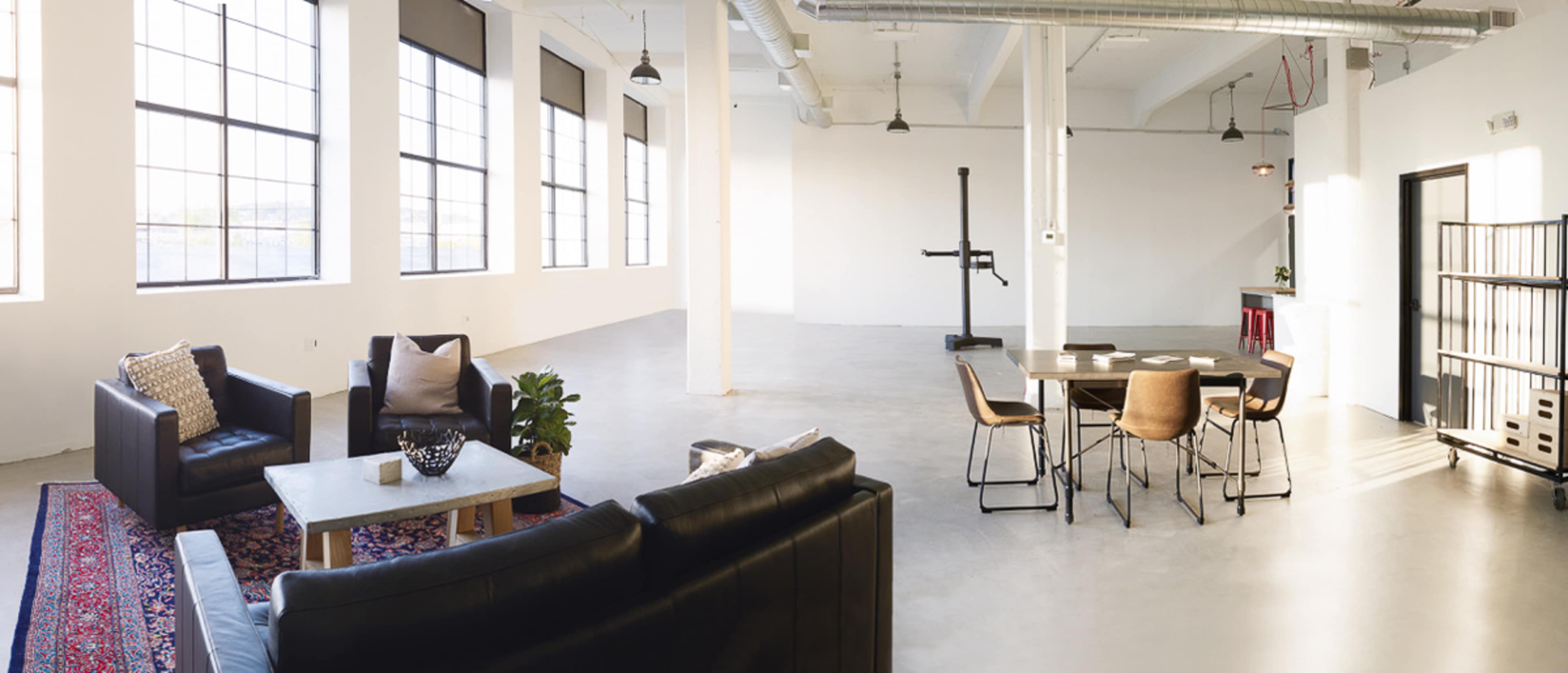 A spacious, modern interior with a seating area featuring black couches, a coffee table, and a dining table set for six, illuminated by large windows.