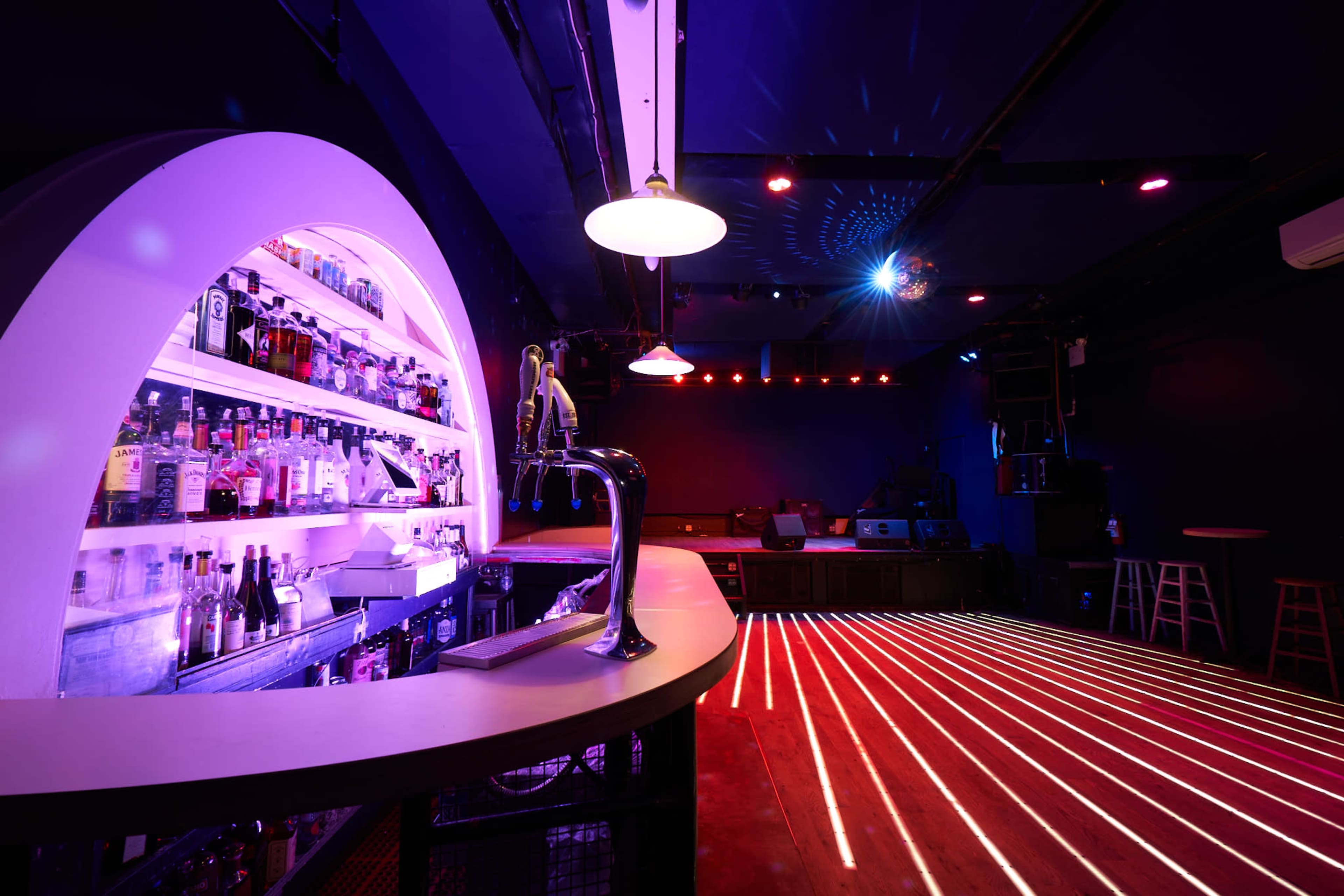 A dimly lit bar with a curved backlit shelf displaying a variety of bottles and a counter with an illuminated surface, accented by colorful lighting effects on the walls and floor.