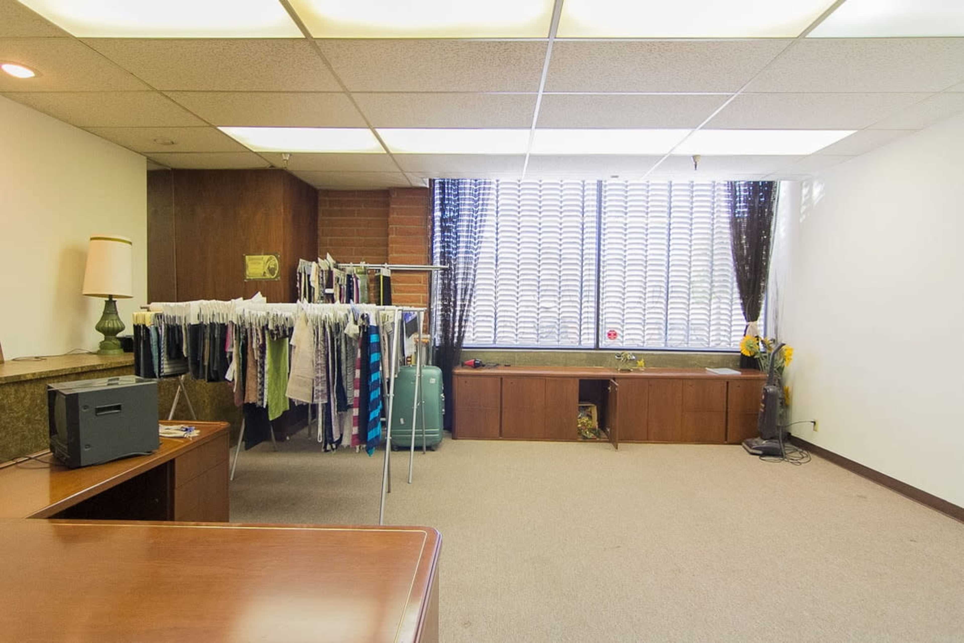 The image shows a spacious room with a clothing rack on one side, a desk and a lamp on another, and a large window with blinds partially drawn.