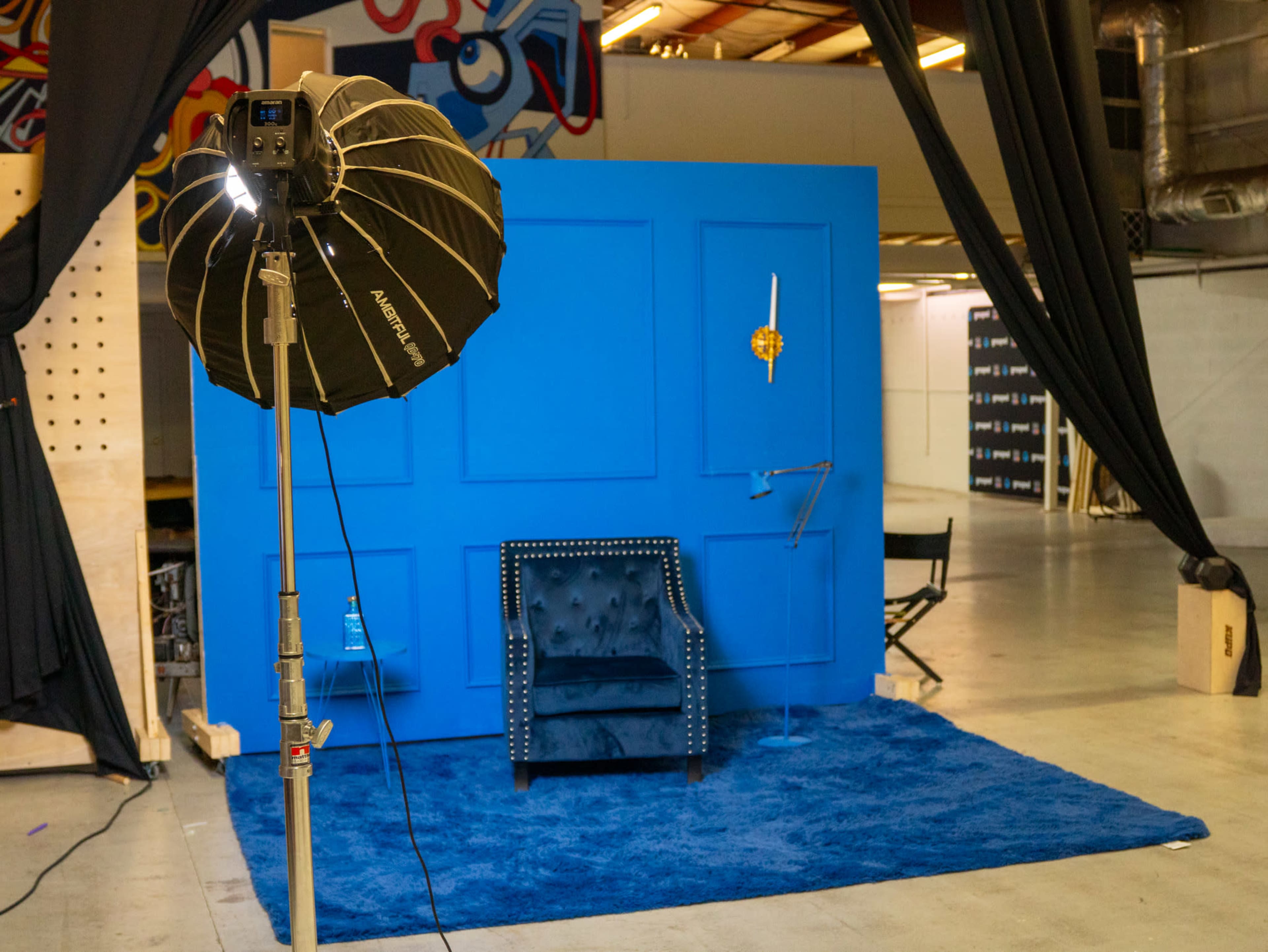 A photo studio setup features a blue backdrop with a chair and a light source on a stand, placed on a blue rug in a spacious area.