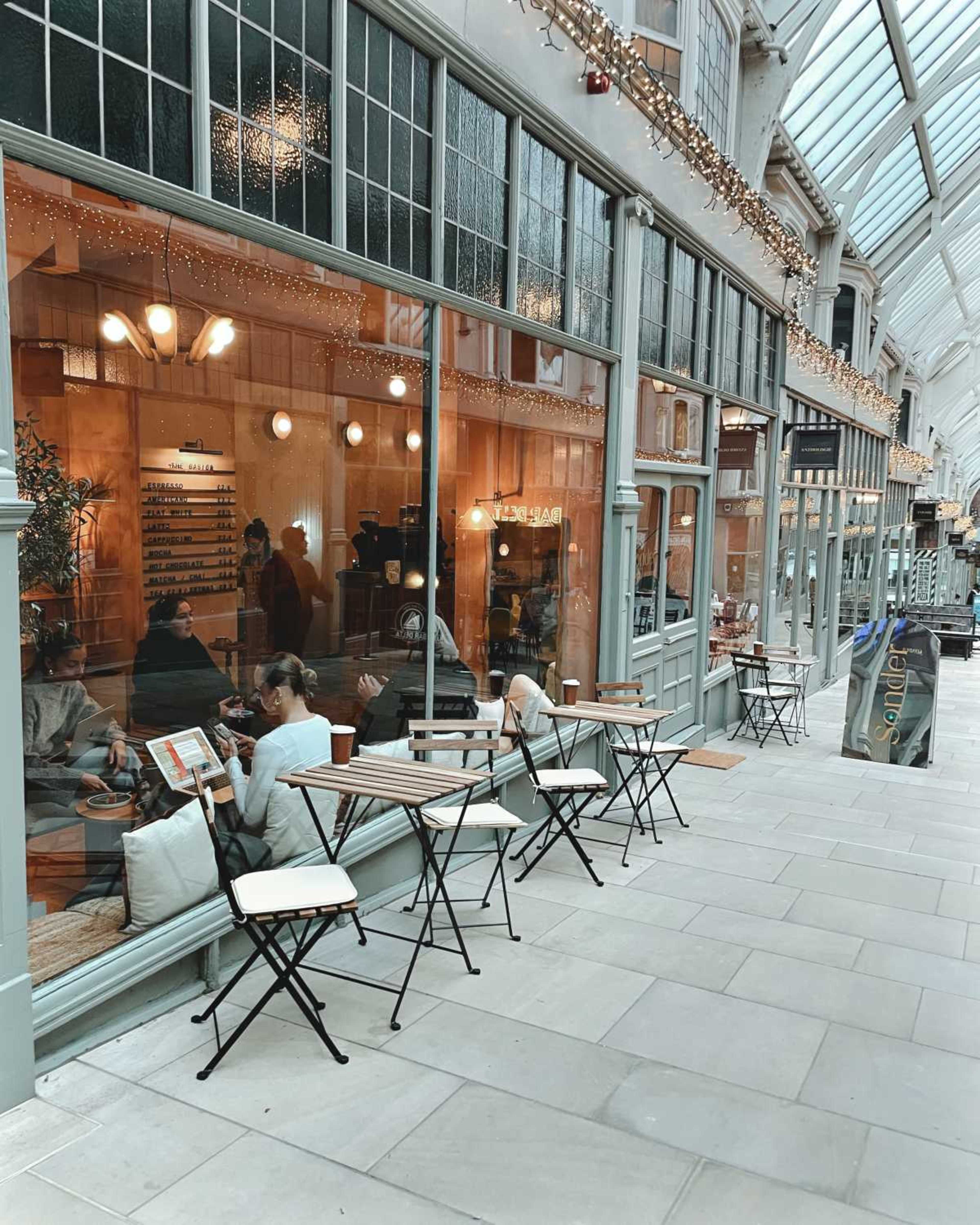 A calm café with outdoor seating is located in a glass-covered arcade, featuring large windows that allow light into the space.