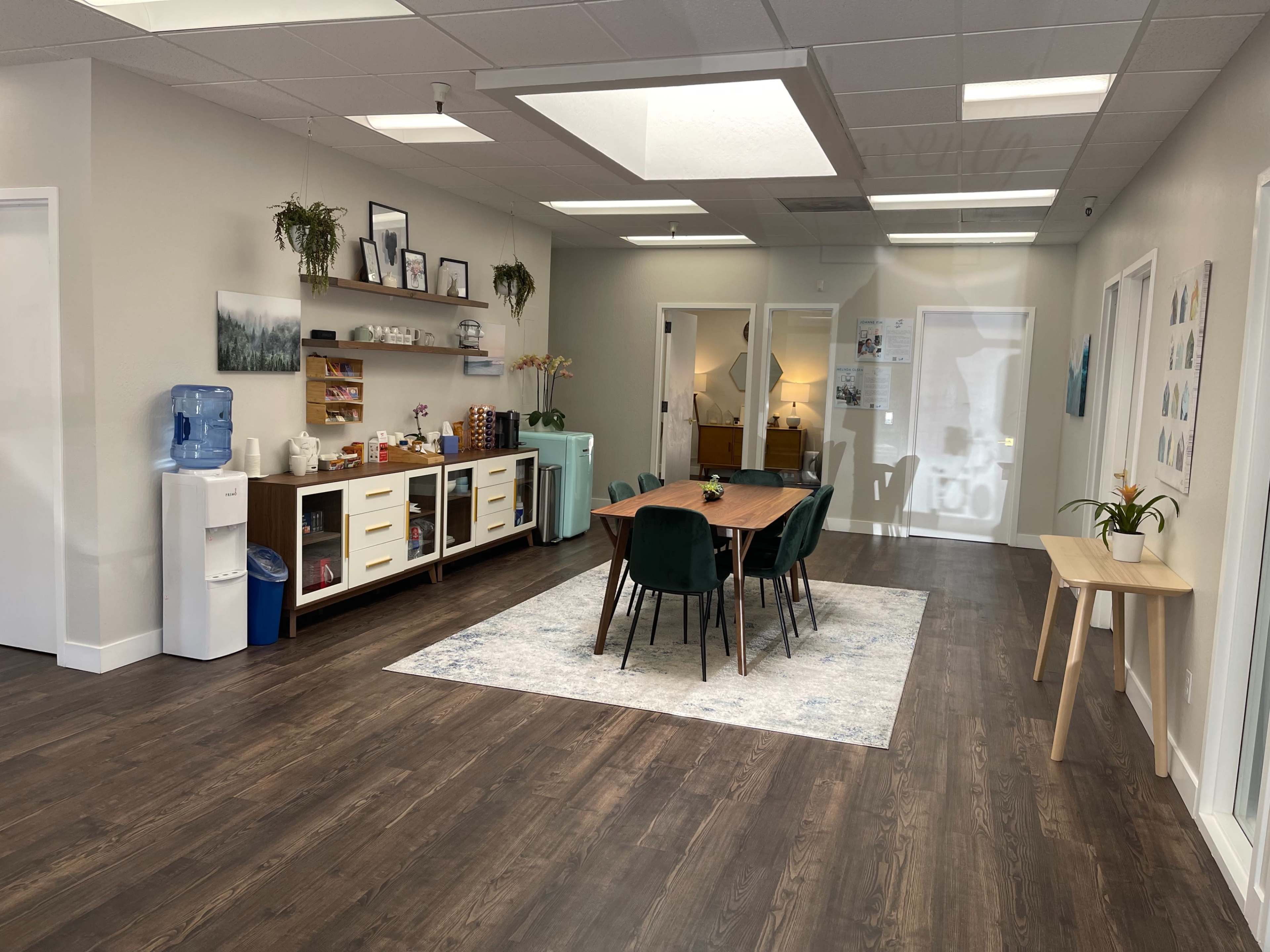 A spacious office interior features a wooden dining table surrounded by green chairs, along with a water cooler and shelves against the walls.