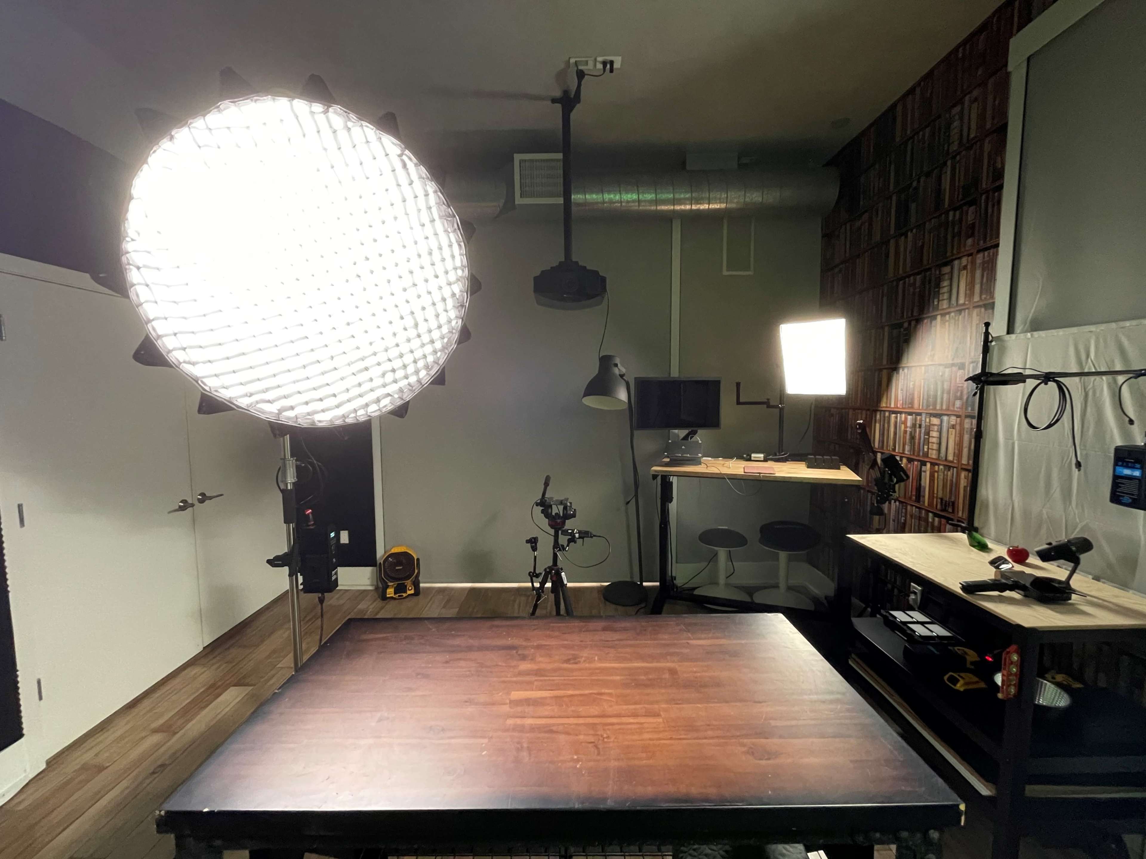 The image shows a photography studio setup with a large circular softbox, a high stool, a camera on a tripod, and various lighting equipment arranged around a wooden table.
