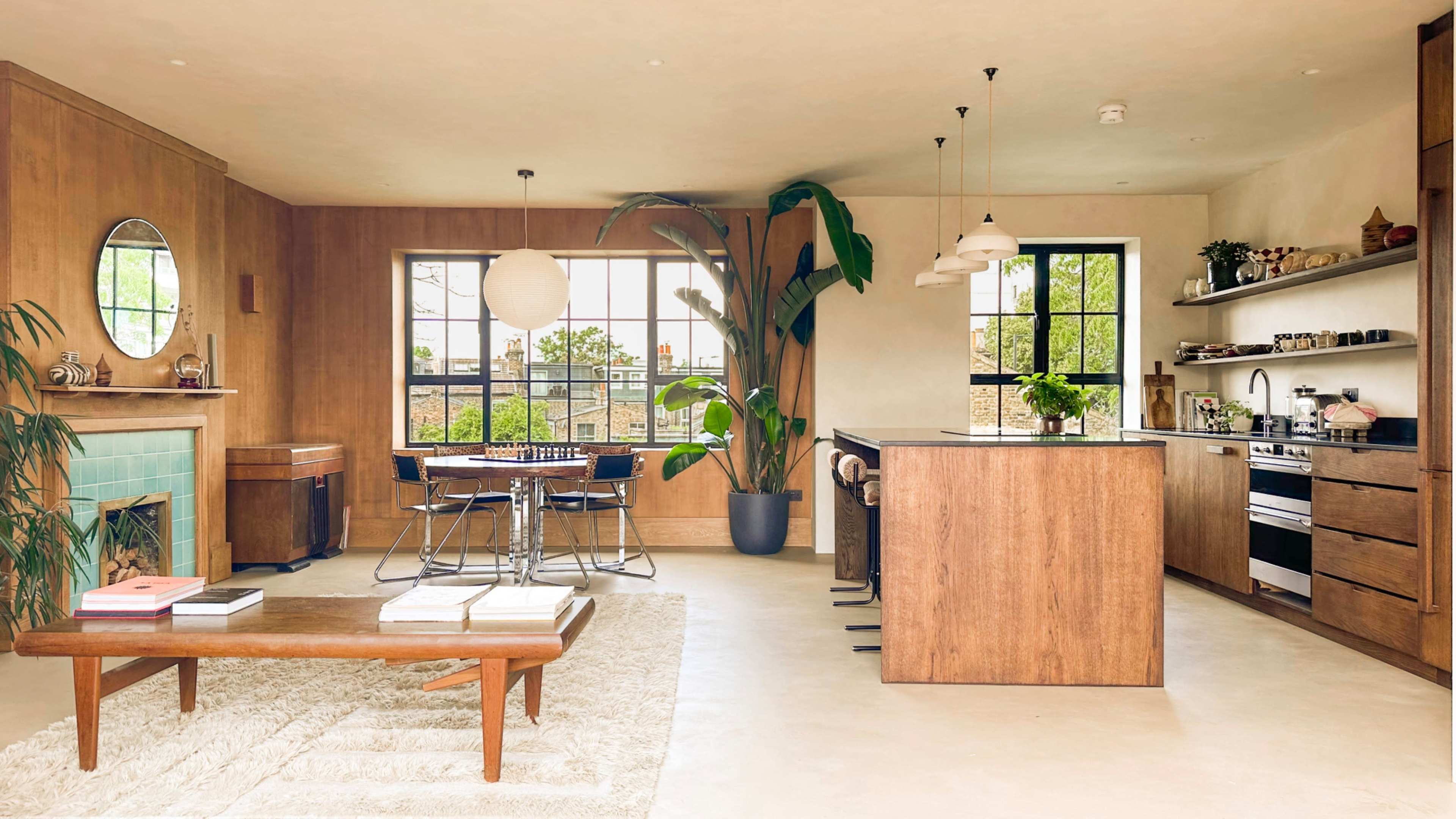The image shows a modern kitchen and living area with wooden finishes, a large window, and a central island, complemented by a plant and minimal furniture.