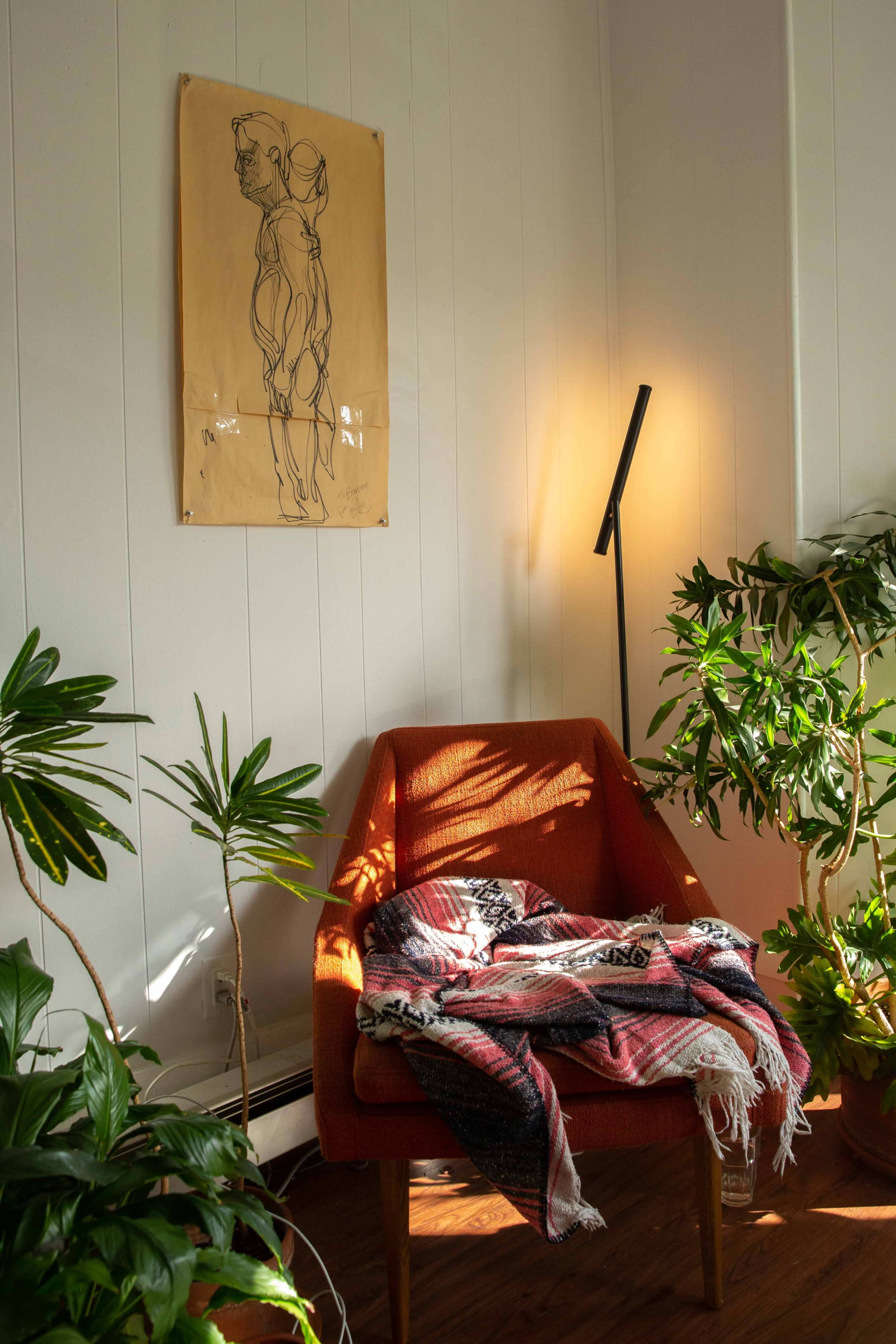 A modern orange armchair sits beside a lamp and is draped with a patterned blanket, surrounded by several green plants and a wall featuring a line drawing.