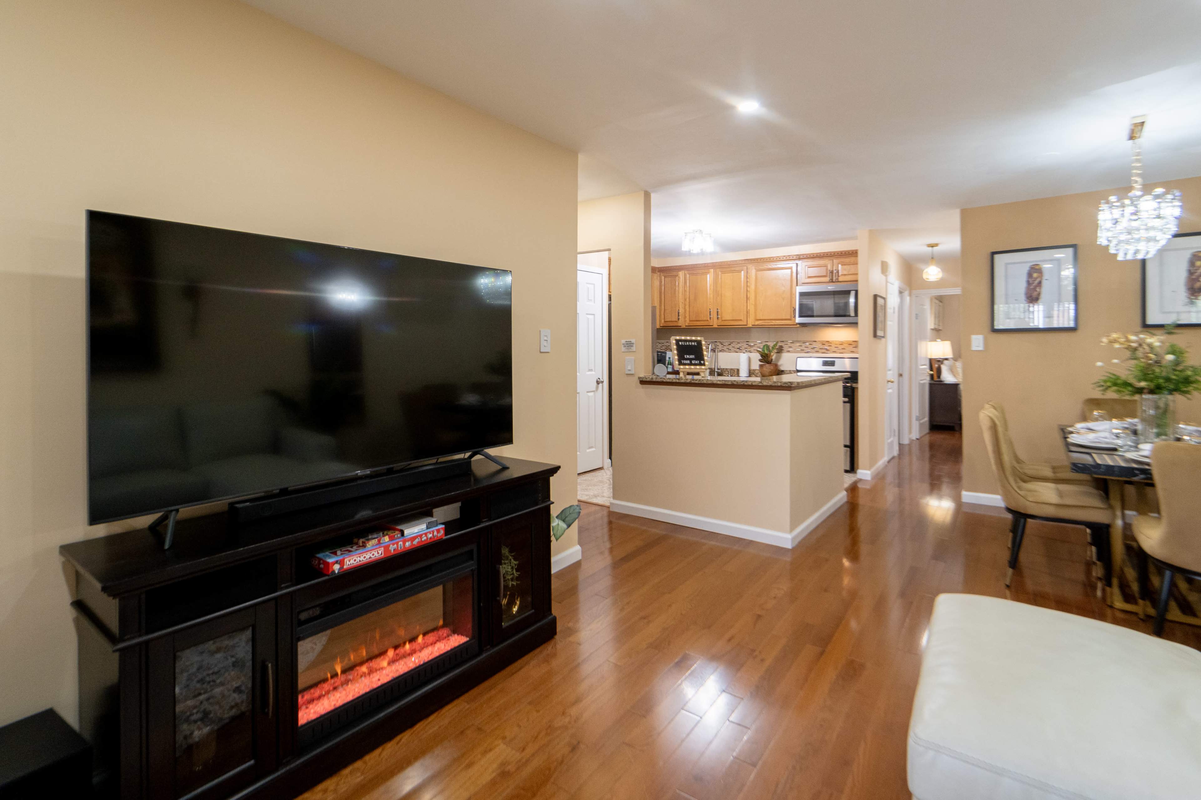 The image shows a living space with a large television mounted on a cabinet, a cozy dining area in the background, and a well-appointed kitchen visible through an open layout.
