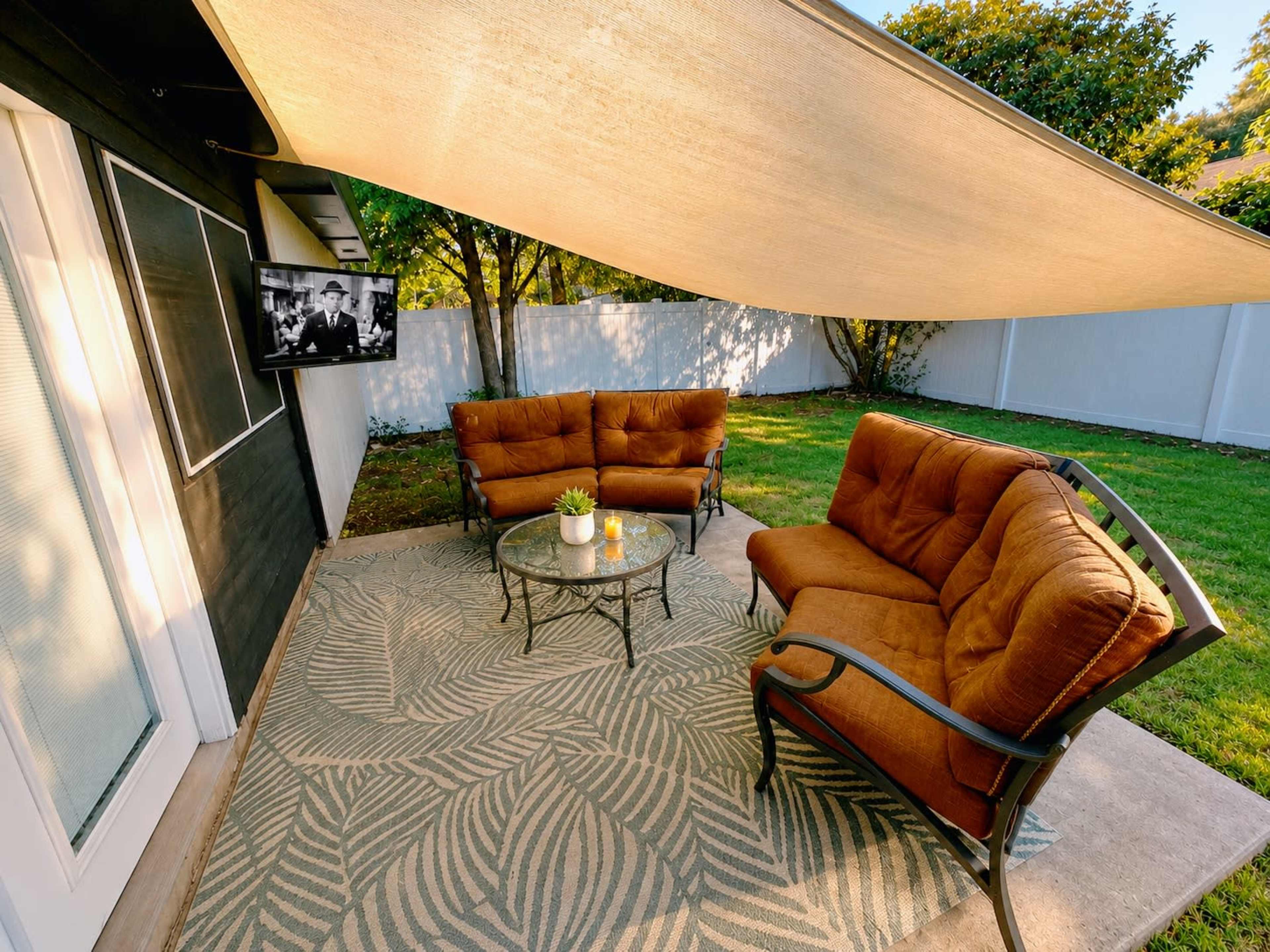 A shaded outdoor seating area features a glass table and upholstered chairs on a patterned rug, with a television mounted on the wall.