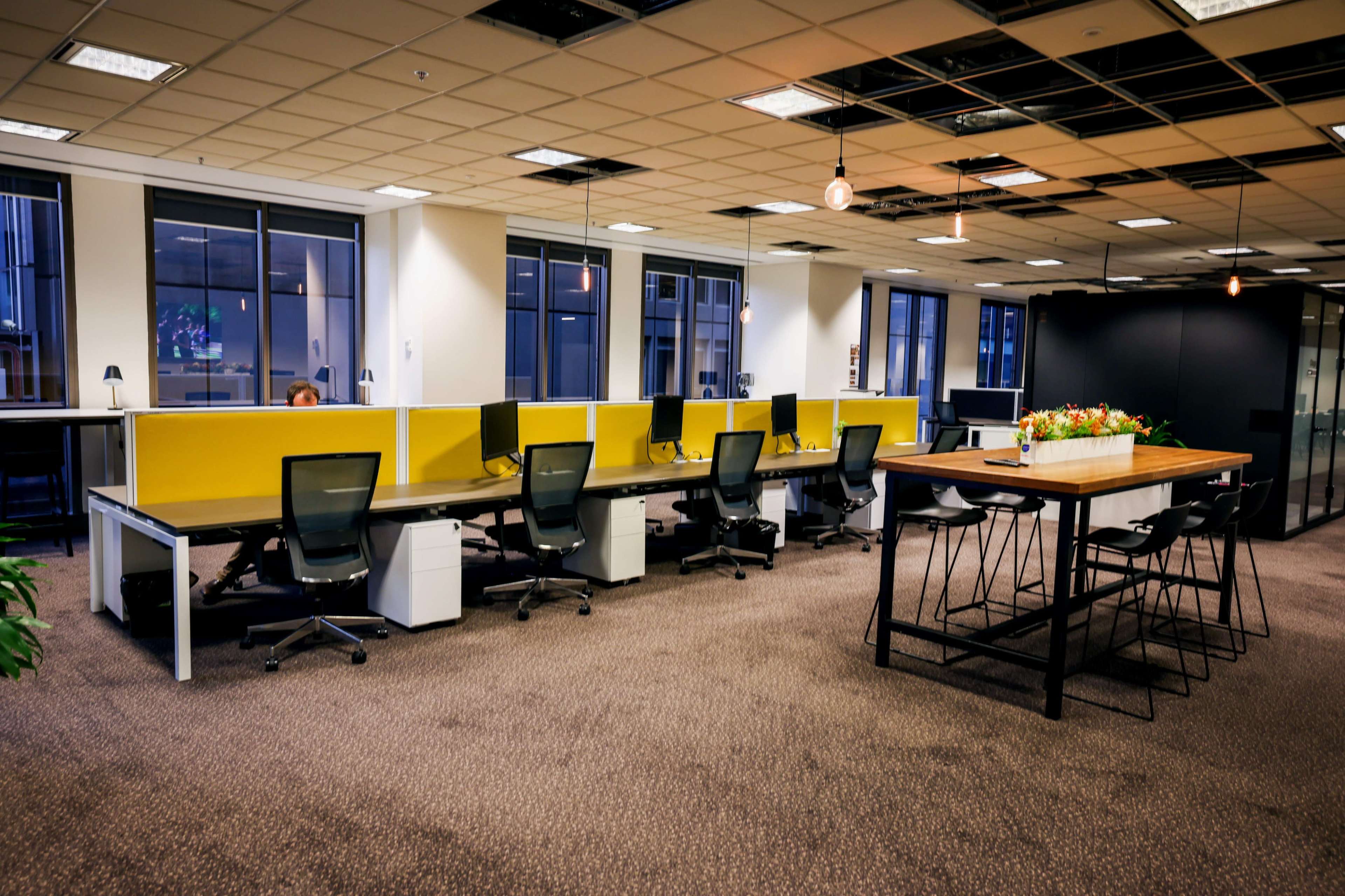 The image shows a modern office space featuring several workstations with computers and a communal table surrounded by chairs.
