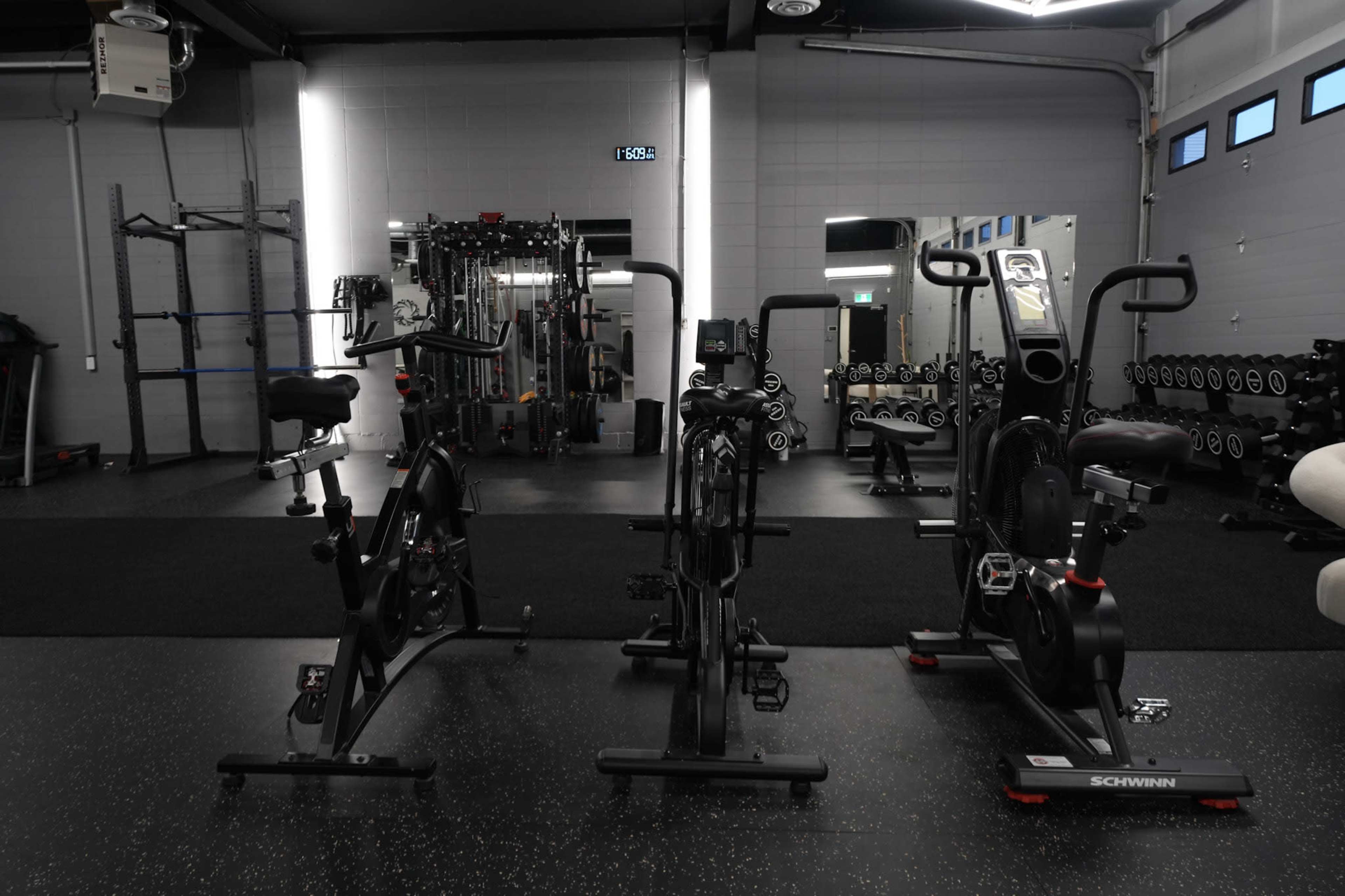 The image shows a gym interior featuring three exercise bikes positioned in front of various fitness equipment, including weights and a cable machine.