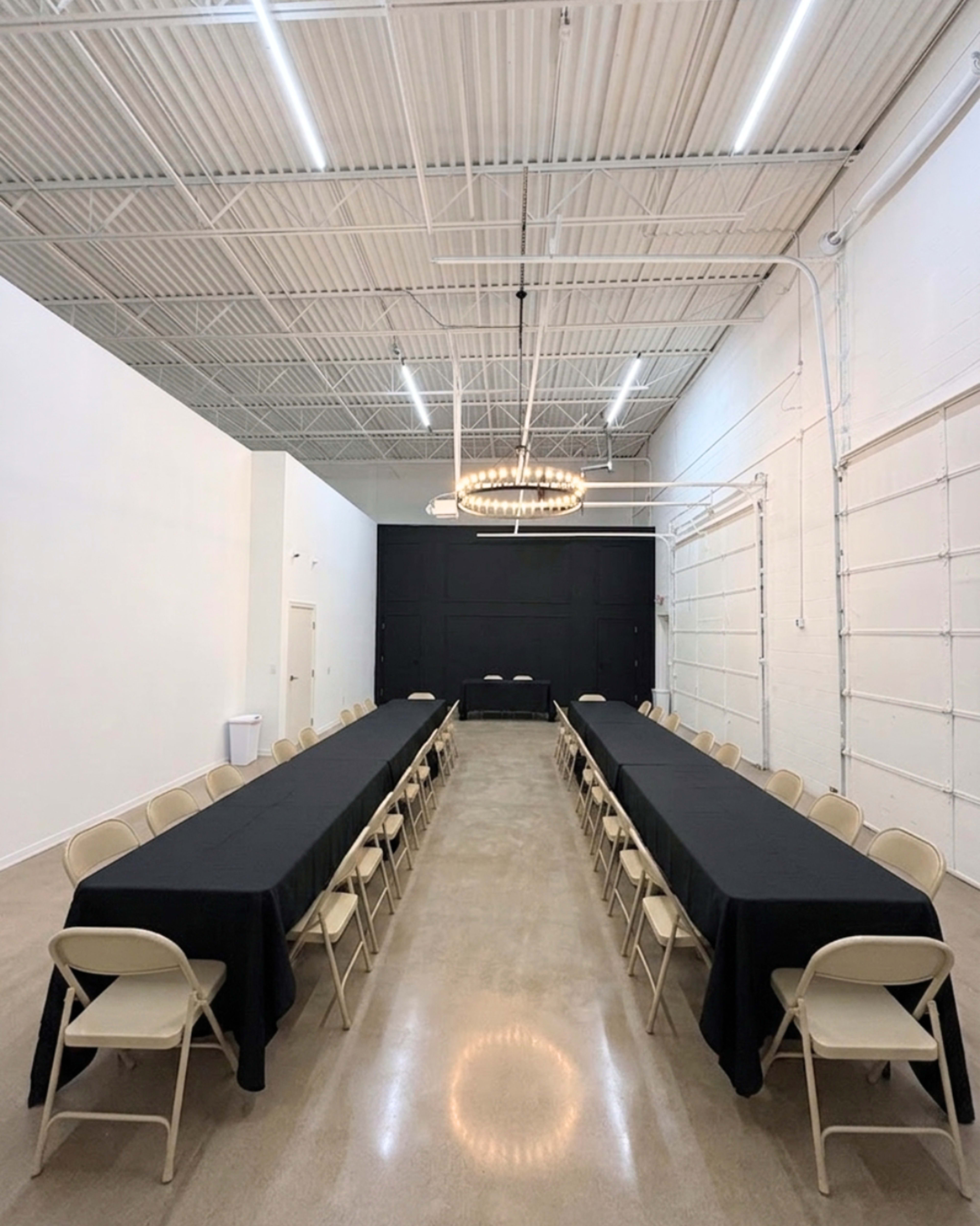 The image shows a large, empty banquet hall with multiple rectangular tables arranged in two rows, covered with black tablecloths, and a chandelier hanging from the ceiling.