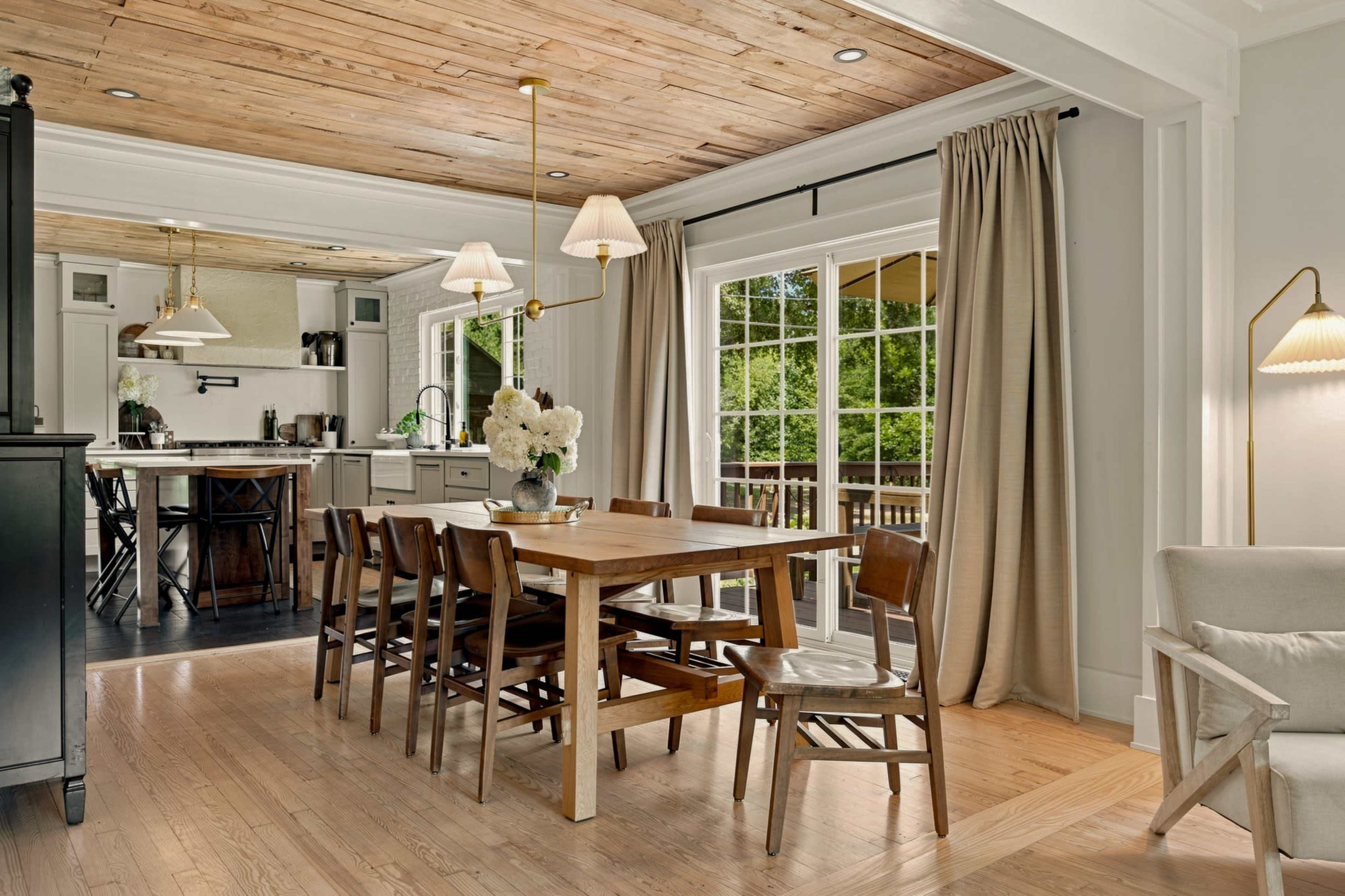 A wooden dining table with chairs occupies the center of a well-lit room featuring a ceiling with wooden planks and large glass doors leading to a deck.
