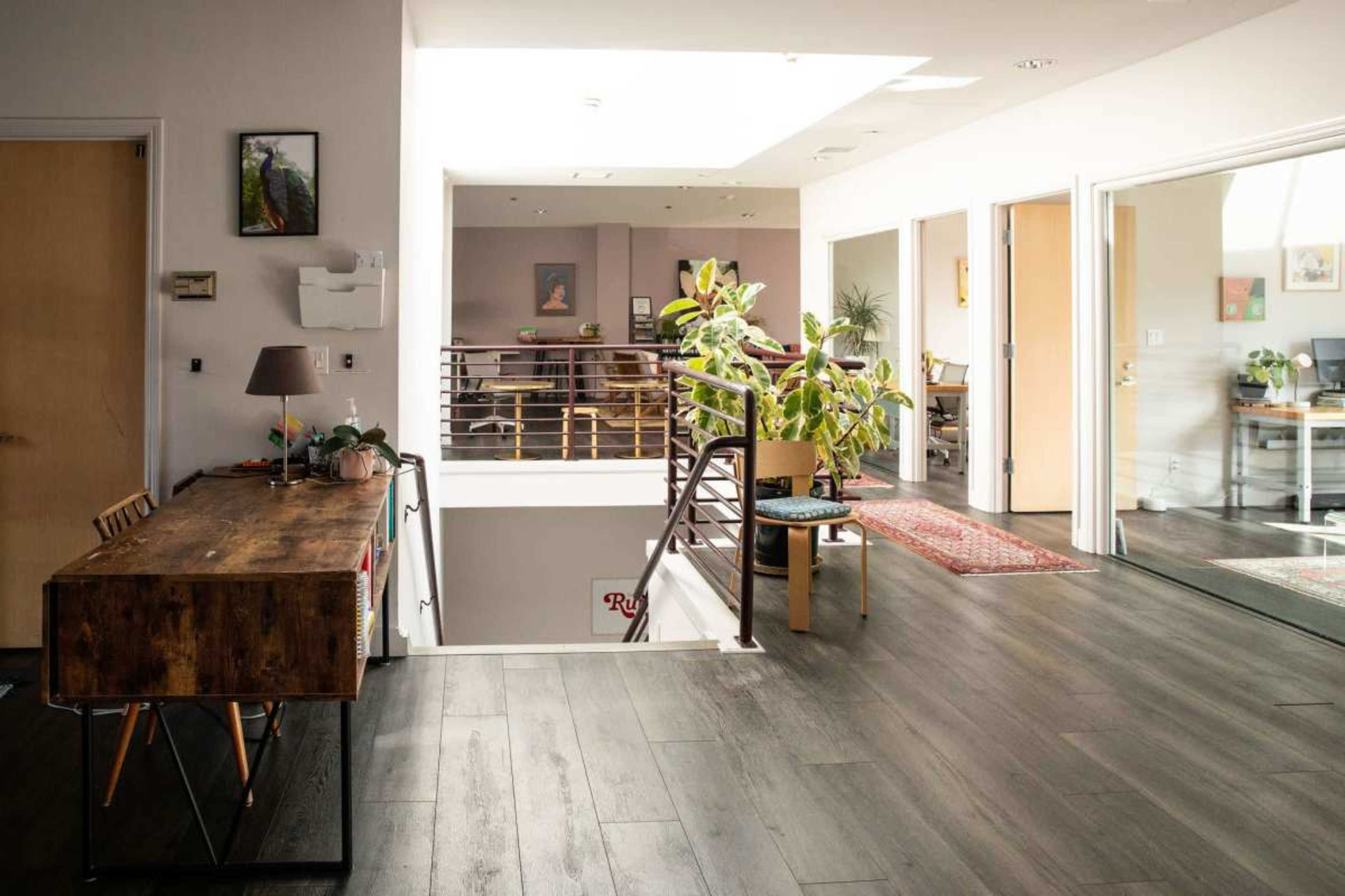 The image shows a modern, open-concept interior space with a wooden desk, plants, and several rooms visible through glass doors.