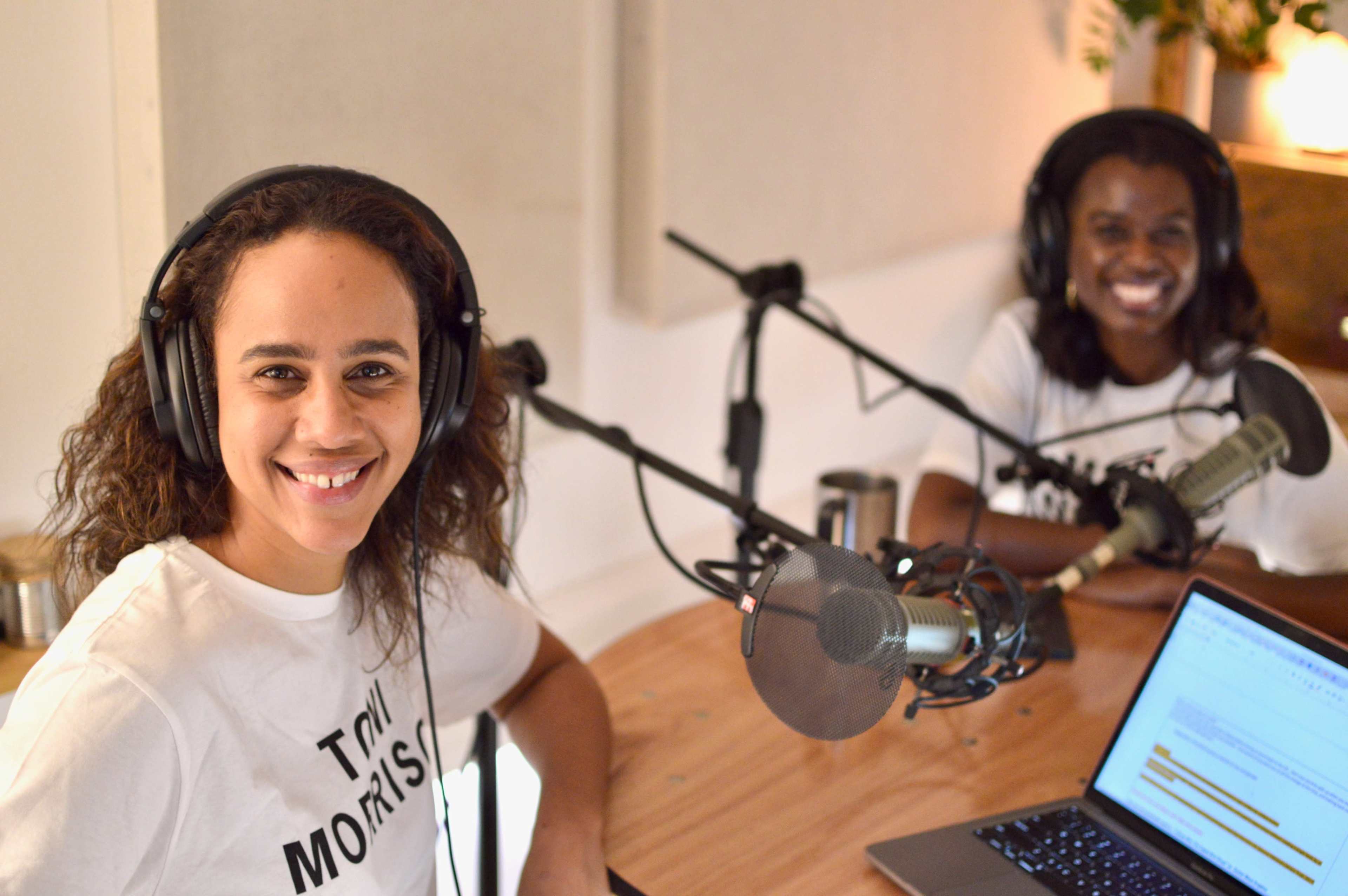 Two women wearing headsets sit at a table with microphones, laptops, and a cozy studio background.