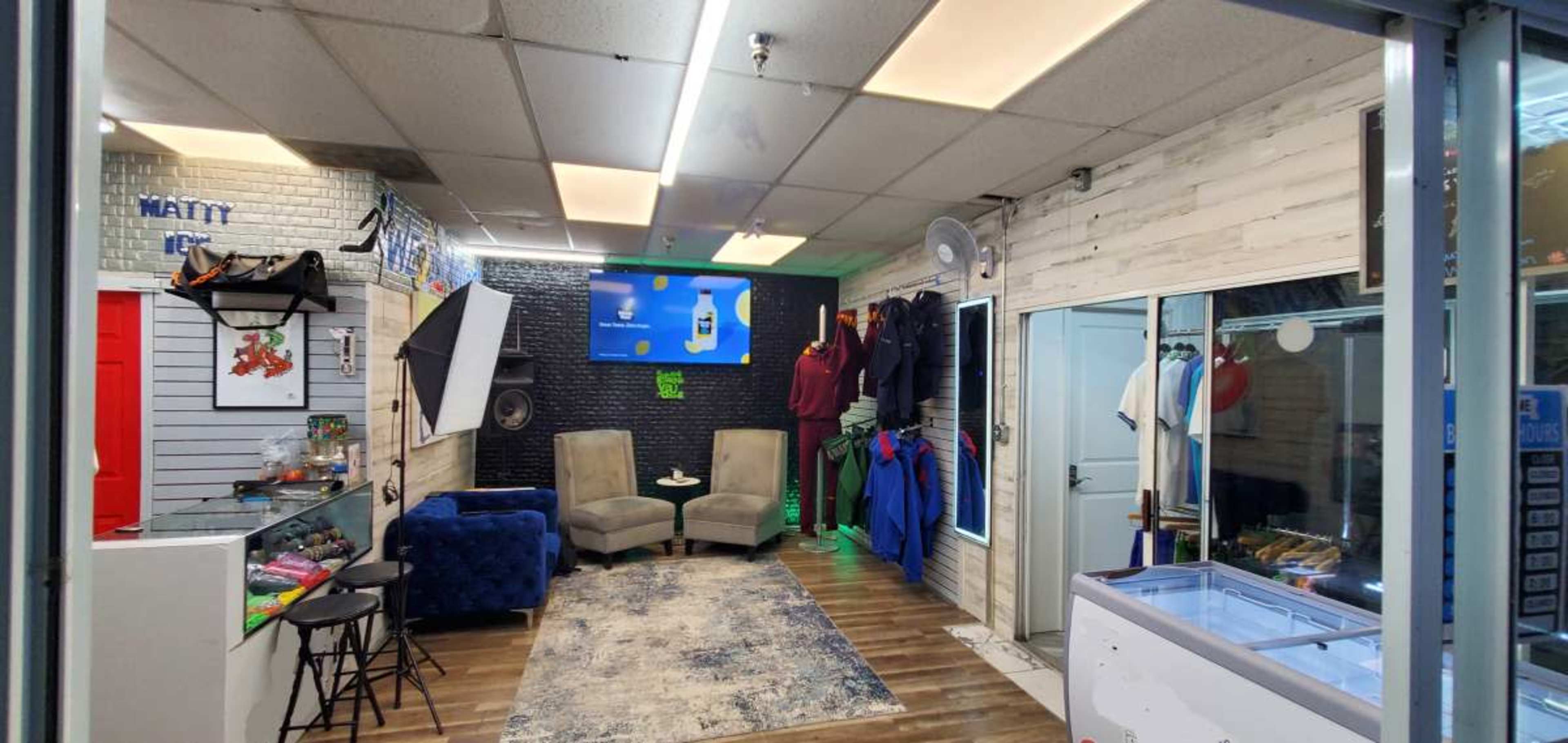 The image shows an indoor retail space with two armchairs, a display area on the left, clothing hanging on the wall, and a freezer in the right corner.