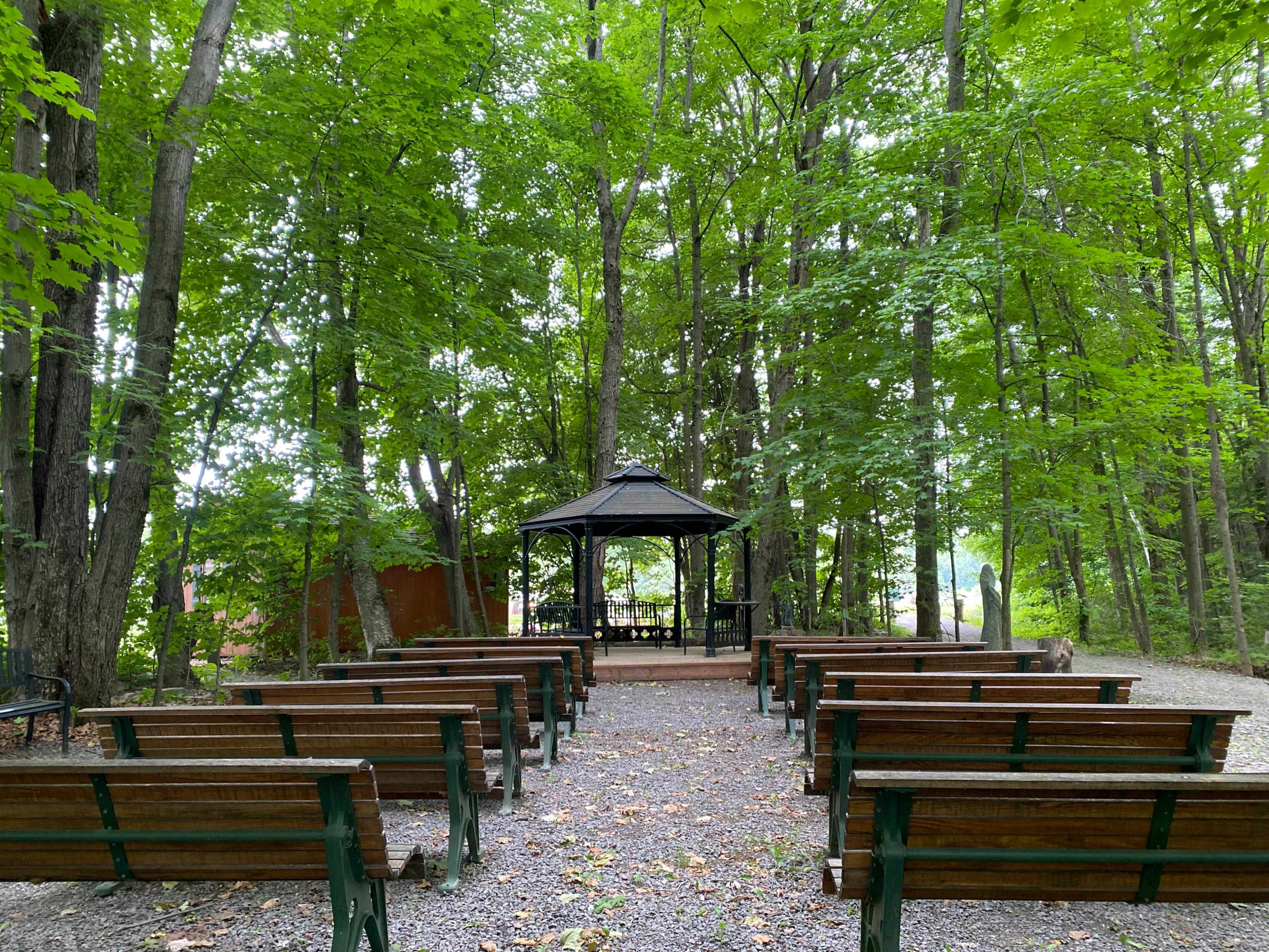 A wooded area features a gazebo at the end of a path lined with benches facing it.