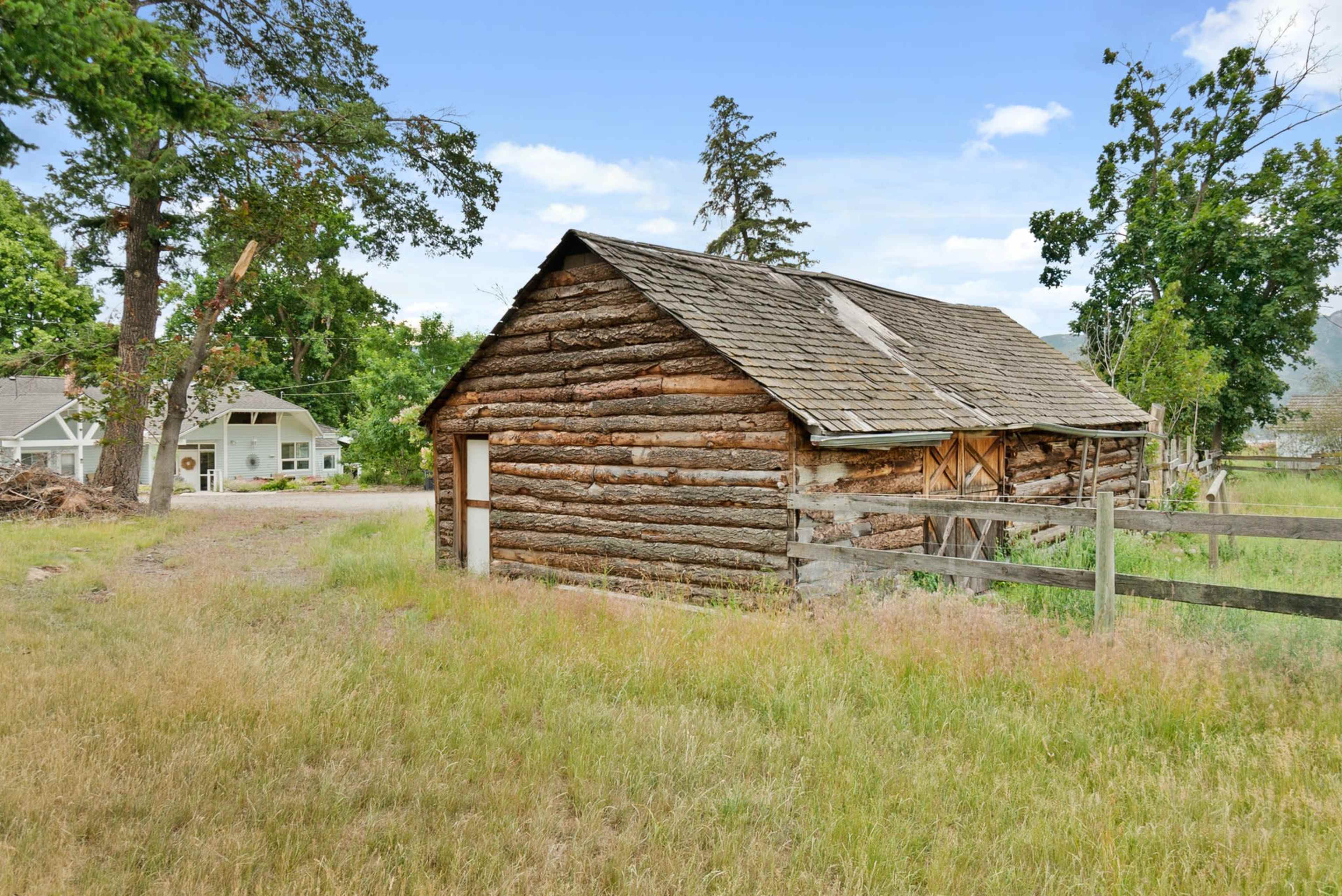 A weathered log cabin sits beside a grassy area, with a white house and trees visible in the background.
