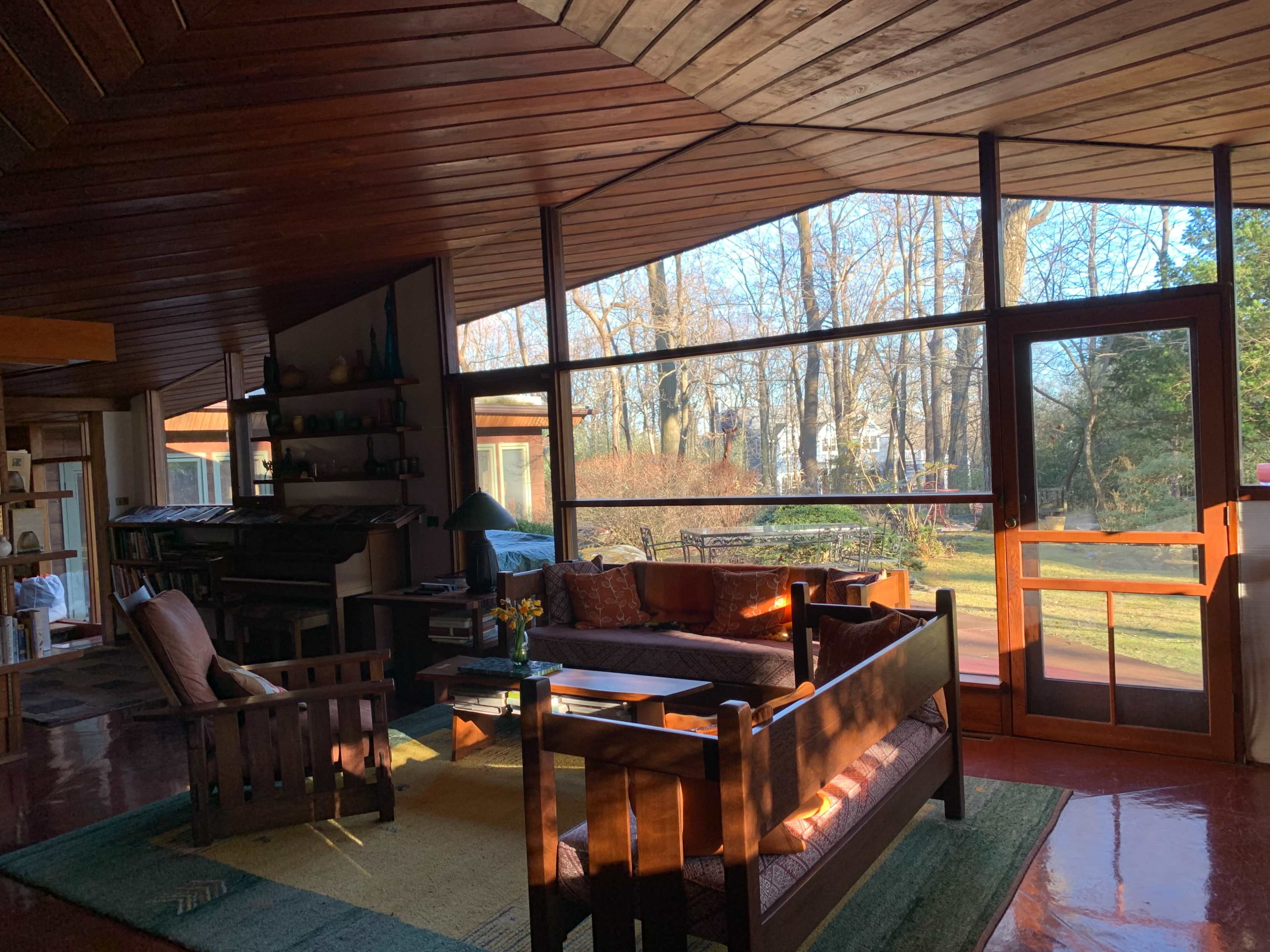 The image shows a spacious living room with wooden walls and ceiling, filled with natural light and featuring a large glass window that opens to a view of trees outside.