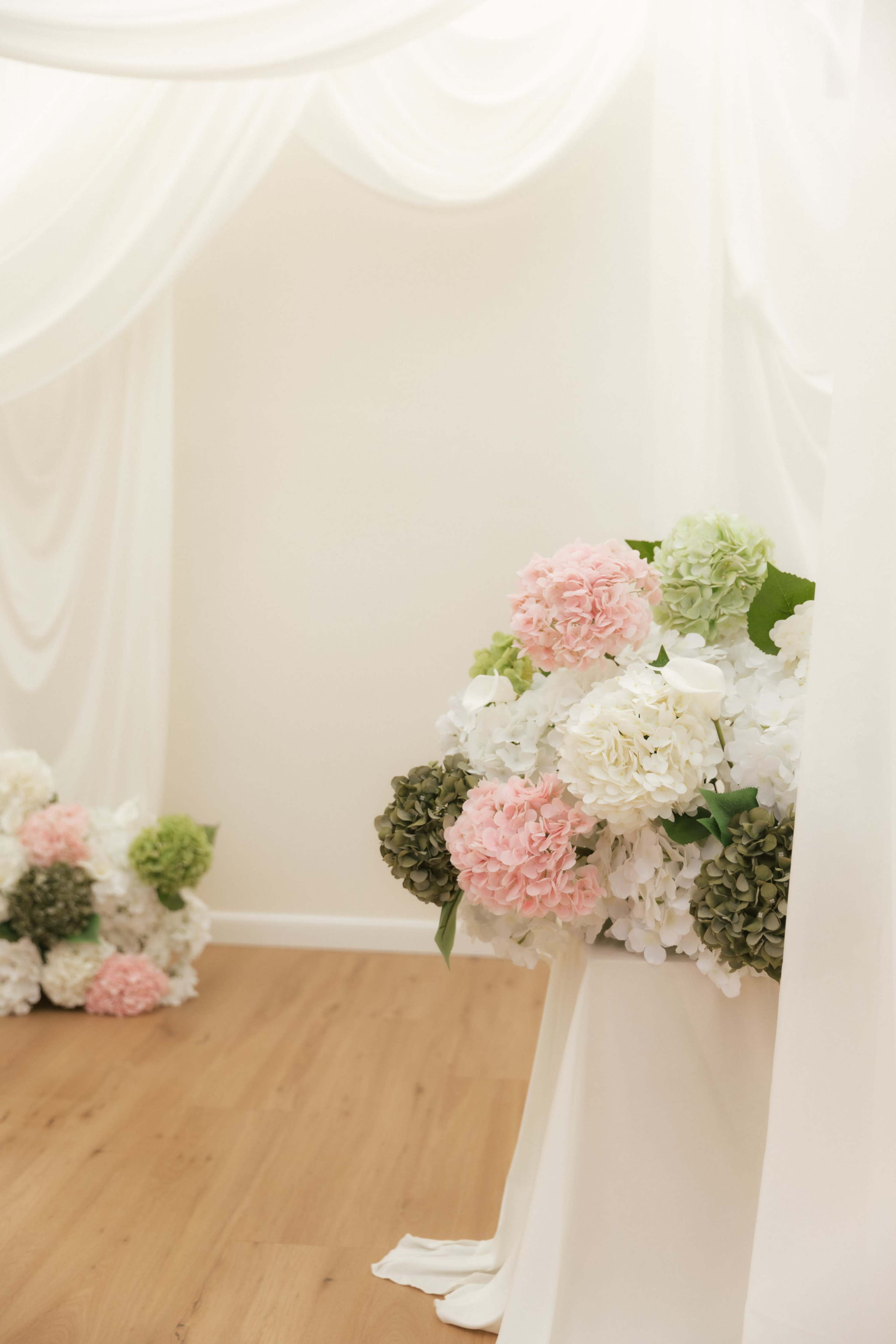 A bouquet of pink, white, and green artificial flowers is arranged near a draped fabric backdrop in a softly lit room.